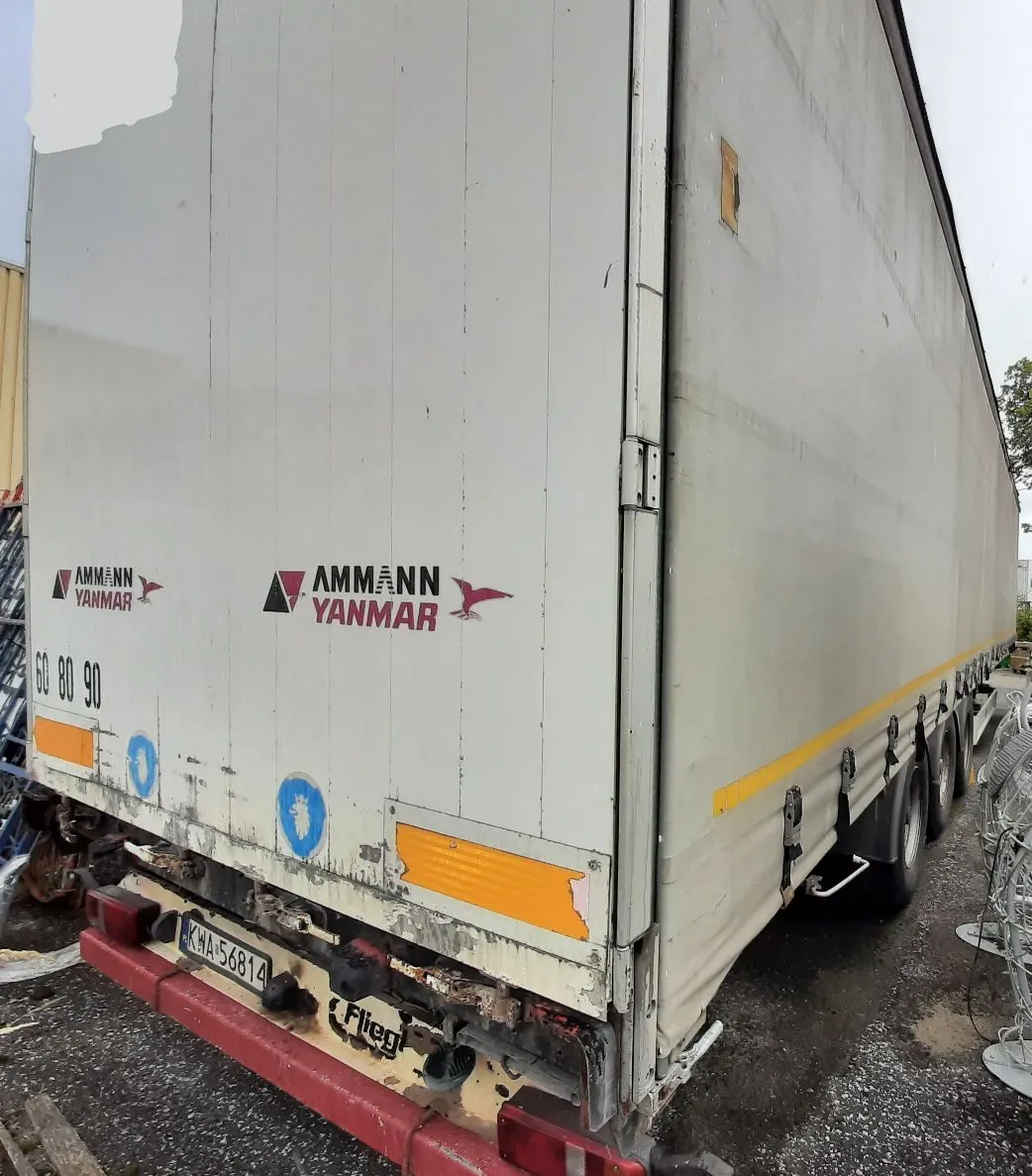 Zdjęcie przedstawiające tylną i boczną część białej naczepy ciężarówki. Na boku naczepy widnieje czerwono-czarny napis „AMMANN YANMAR” wraz z logo przedstawiającym stylizowanego ptaka. W dolnej części naczepy znajduje się poziomy, żółty odblaskowy pas. Na zderzaku widoczna jest tablica rejestracyjna o numerze „KWA 56814” oraz napis „Frigo”. Konstrukcja naczepy wykazuje oznaki zużycia i zabrudzeń.
