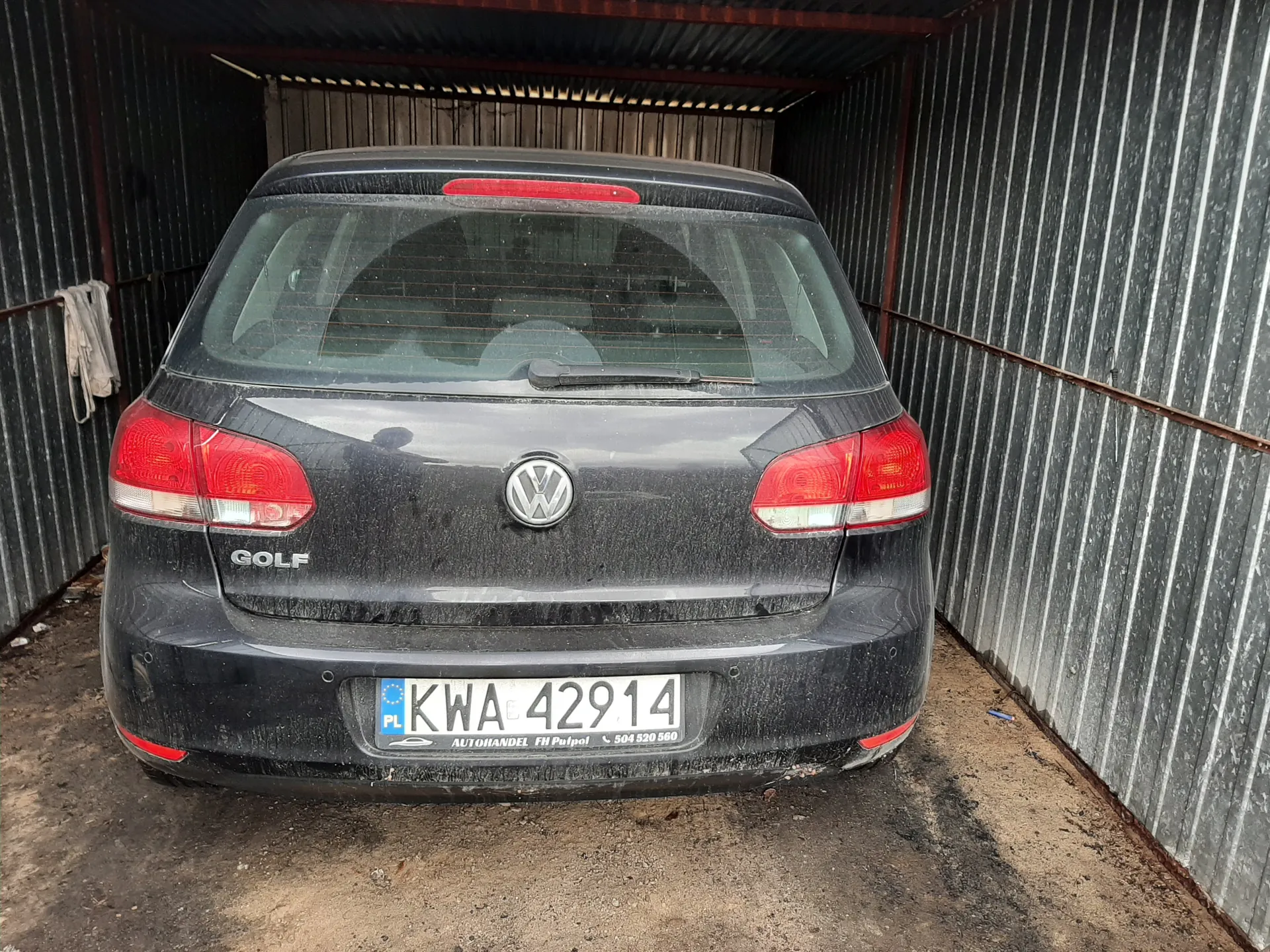 Tył czarnego samochodu Volkswagen Golf zaparkowanego w metalowym garażu. Na bagażniku widoczny napis "GOLF" oraz logo VW. Tablica rejestracyjna z polskim kodem "PL" i numerem "KWA 42914". Poniżej tablicy napis "AUTOHANDEL FH Pulpot" oraz numer telefonu "504 520 560". Samochód pokryty jest kurzem, a w tle metalowe ściany garażu z wiszącym ręcznikiem po lewej stronie.