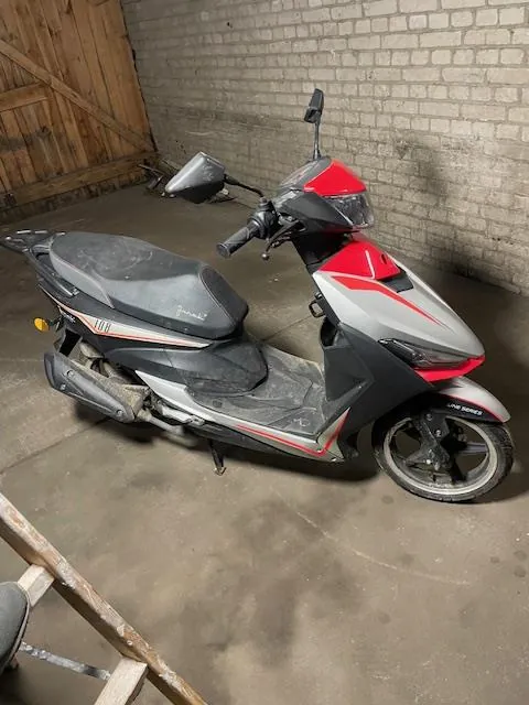 Czerwono-szary skuter z czarnym siedzeniem zaparkowany w pomieszczeniu z betonową podłogą. W tle widoczna ceglana ściana oraz drewniane drzwi. Skuter posiada lusterka, reflektor i widoczne elementy konstrukcyjne. Na podłodze widoczne ślady oraz fragment drewnianego obiektu w lewym dolnym rogu.