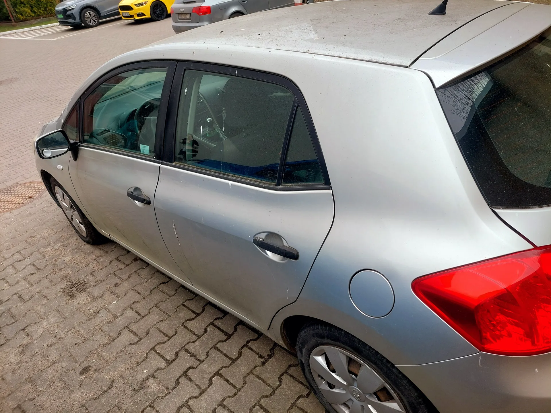 Srebrny samochód osobowy typu hatchback zaparkowany na brukowanym placu parkingowym. Widoczne są ślady uszkodzeń na boku pojazdu oraz czerwone światło tylnego. W tle inne zaparkowane samochody: żółty, szary i srebrny. Podłoga złożona z kamiennych płyt ułożonych w wzór.