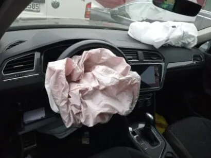 Wnętrze samochodu z rozwiniętymi poduszkami powietrznymi. Na kierownicy widoczna różowa poduszka powietrzna, a na desce rozdzielczej po stronie pasażera biała poduszka. W tle przez przednią szybę widać fragment białego pojazdu z częściowo widoczną tablicą rejestracyjną zawierającą cyfry '585'. Wnętrze samochodu ma ciemne elementy, w tym kierownicę, deskę rozdzielczą i centralną konsolę z przyciskami oraz ekranem. Widoczny jest również dźwignia zmiany biegów.