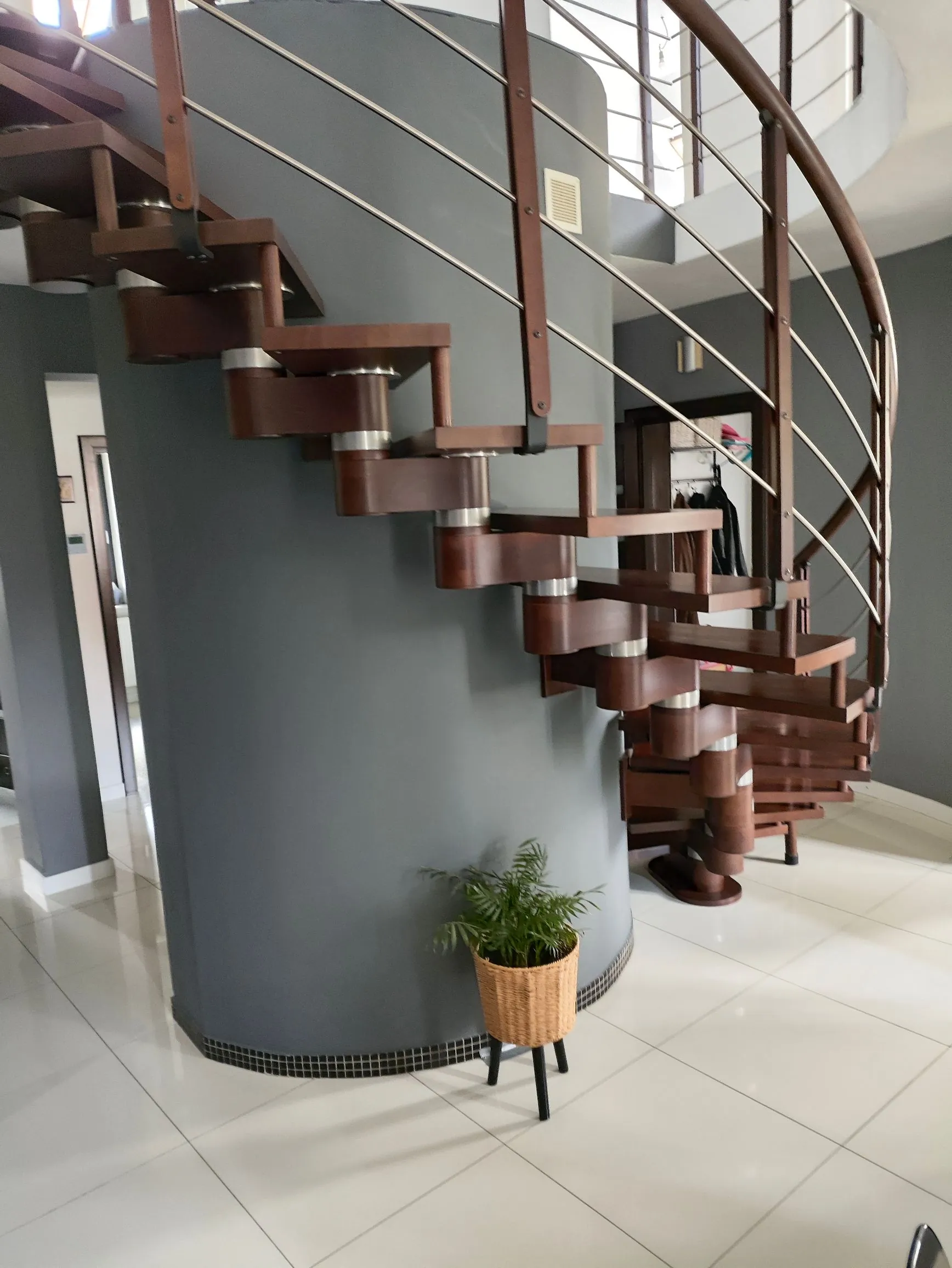 Nowoczesne, spiralne schody z ciemnego drewna z metalowymi elementami konstrukcyjnymi i cienkimi, poziomymi prętami pełniącymi rolę balustrady. Schody owijają się wokół szarej, cylindrycznej ściany. Na białej, błyszczącej podłodze, u podstawy szarej ściany, stoi mała zielona roślina w plecionej doniczce na trzech czarnych nóżkach.