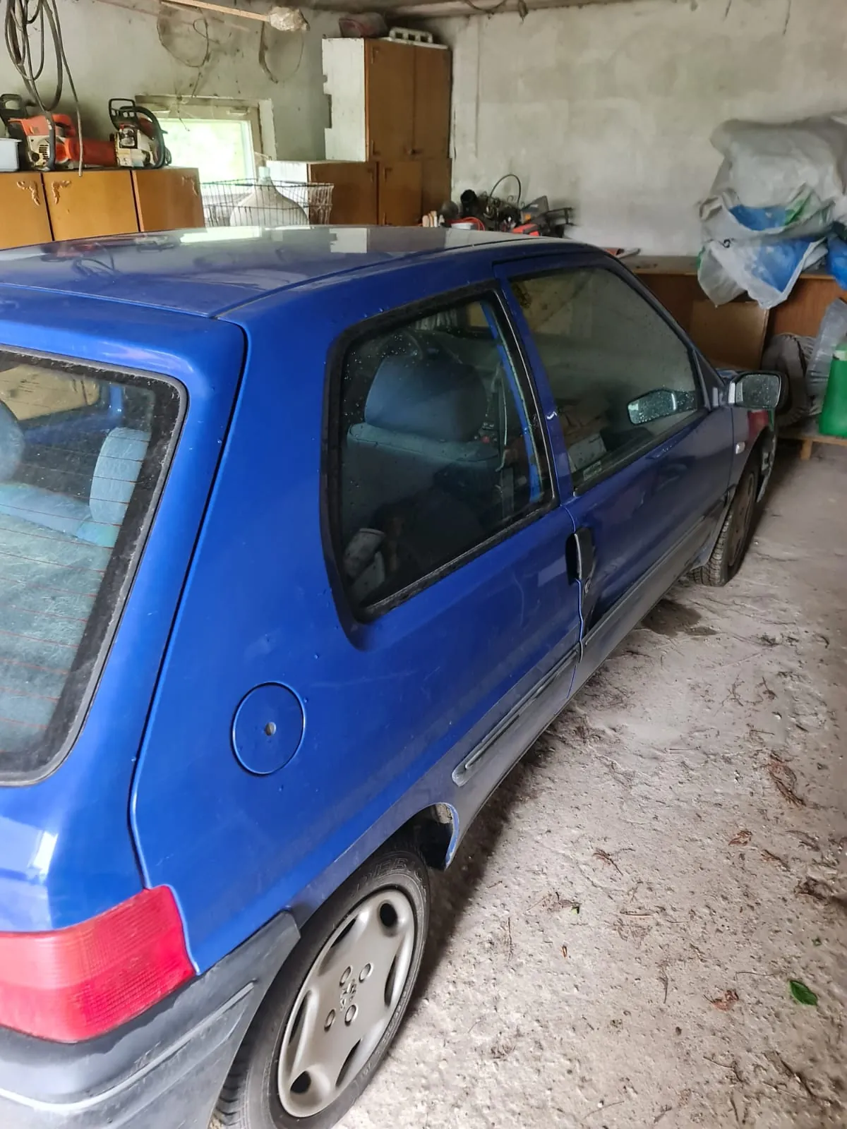Niebieski samochód osobowy typu hatchback zaparkowany w garażu. W tle widoczne betonowe ściany, drewniane szafy, narzędzia i sprzęt na półkach, okno oraz worki i pojemniki po prawej stronie. Podłoga garażu jest brudna, z widocznymi śladami kurzu i śmieci. Samochód ma czerwone światła tylnego, srebrne felgi oraz czarne elementy nadkoli. Wnętrze garażu zawiera również zielony pojemnik i różne przedmioty na półkach.