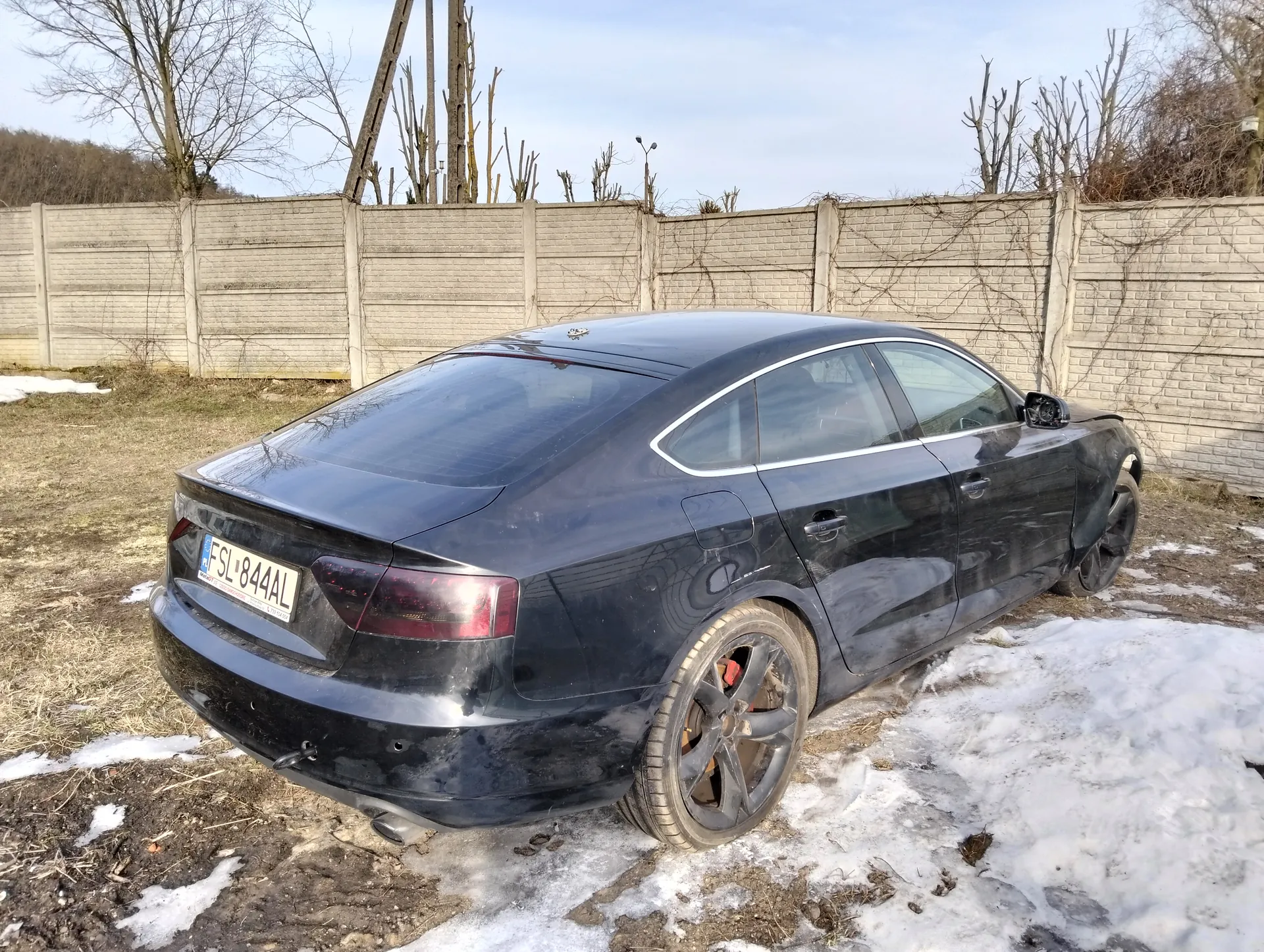 Czarny samochód osobowy typu sedan z tablicą rejestracyjną FSL-844AL zaparkowany na terenie z resztkami śniegu i suchą trawą. W tle betonowy płot, nagie drzewa oraz jasne niebo.