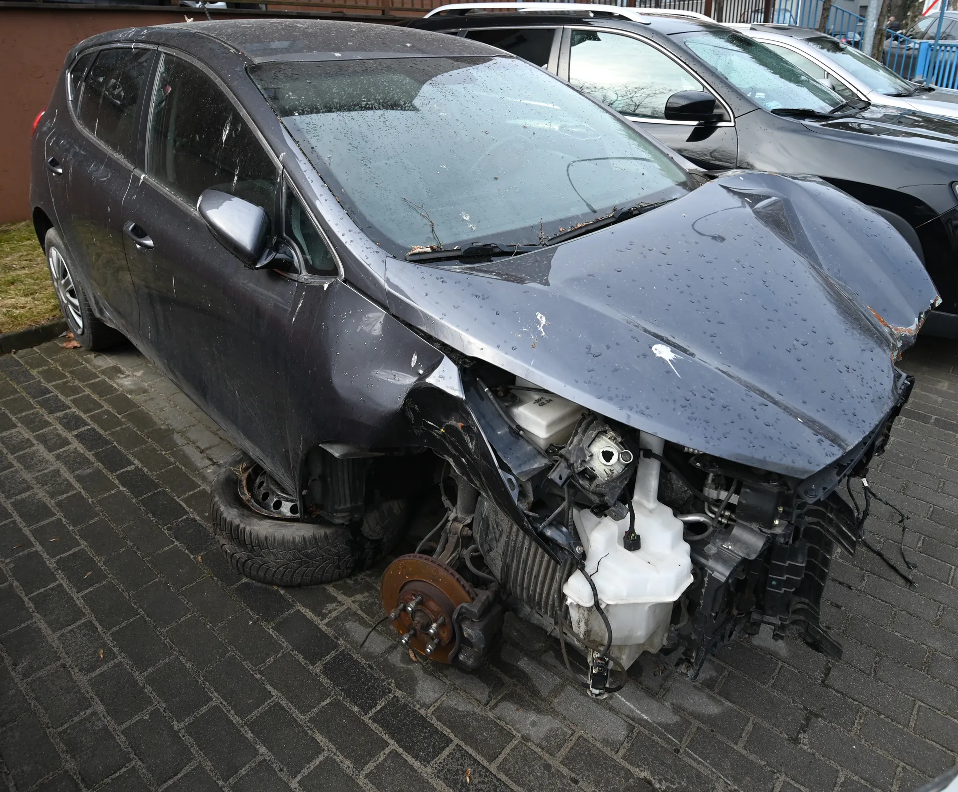 Zniszczony ciemnoszary samochód osobowy z uszkodzonym przodem. Widoczne zdeformowane nadwozie, odsłonięty silnik, brak przedniego koła oraz leżąca obok opona. Samochód stoi na brukowanej powierzchni, w tle inne pojazdy i fragment budynku. Na przedniej szybie krople wody.