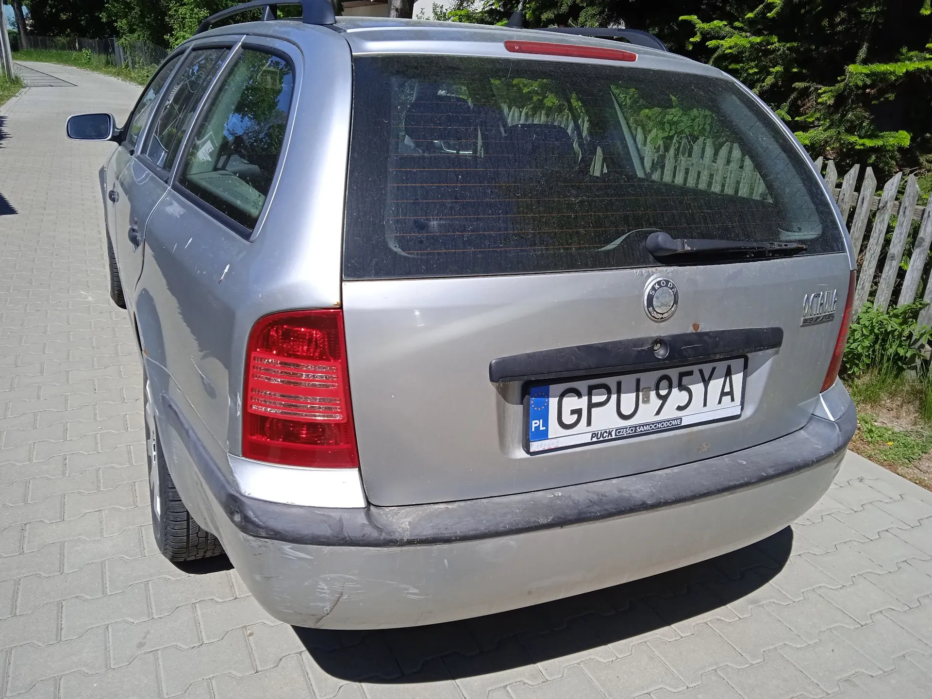 Srebrny samochód osobowy typu kombi zaparkowany na kostce brukowej. Widoczny jest tył pojazdu z tablicą rejestracyjną o numerze GPU 95YA. Na tablicy znajduje się oznaczenie PL oraz napis 'PUCK GIEŁDA SAMOCHODÓW'. Samochód ma czarne relingi dachowe i czarny zderzak. W tle widoczny jest drewniany płot oraz zielone drzewa i krzewy.