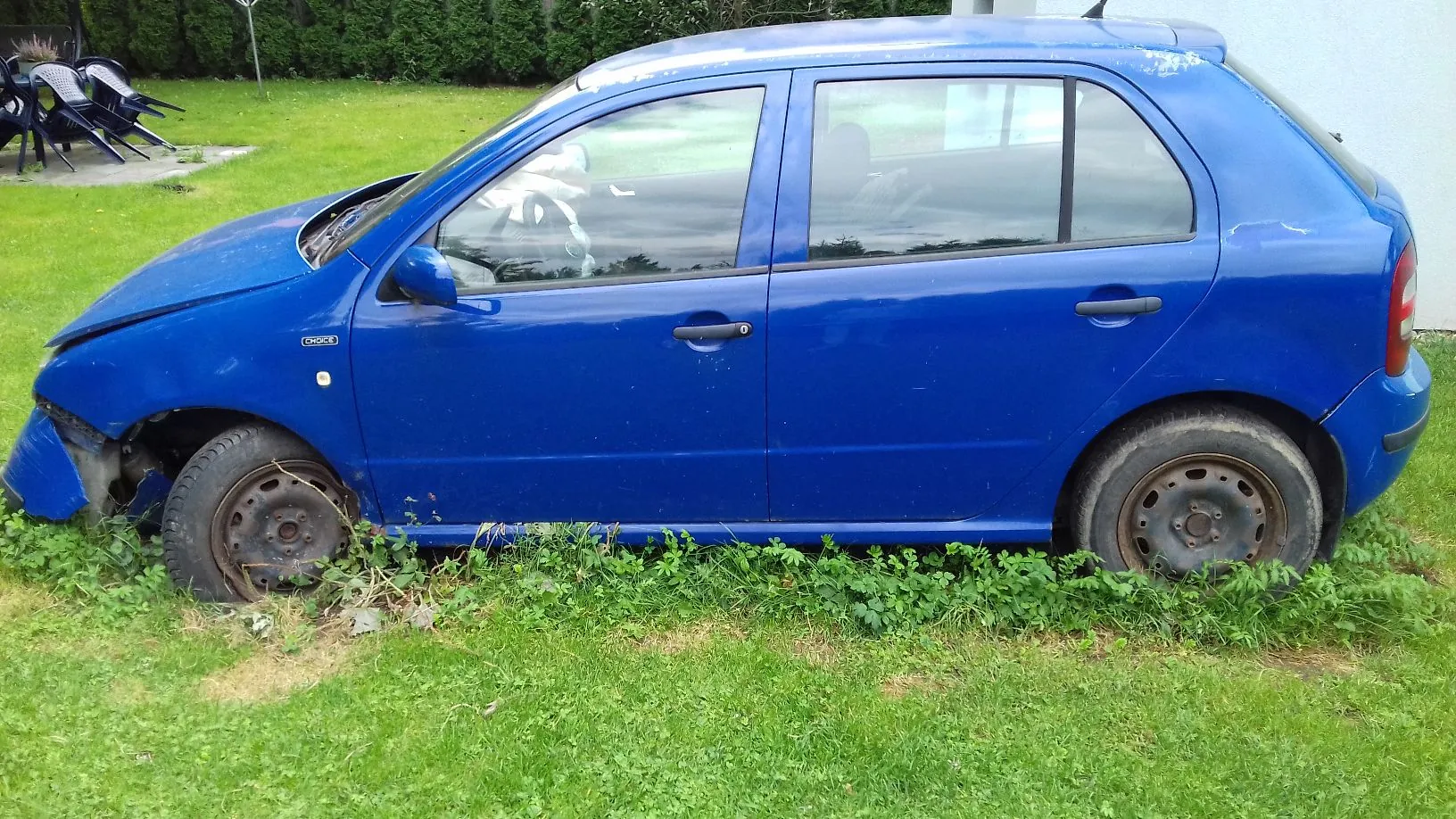 Niebieski samochód osobowy typu hatchback zaparkowany na trawie. Pojazd jest uszkodzony w przedniej części po stronie kierowcy, brakuje przedniego lewego błotnika i reflektora, a zderzak jest częściowo oderwany. Samochód stoi na trawniku, a w dolnej części nadwozia widoczne są kępy zielonych roślin. W tle widać zielony trawnik i żywopłot.