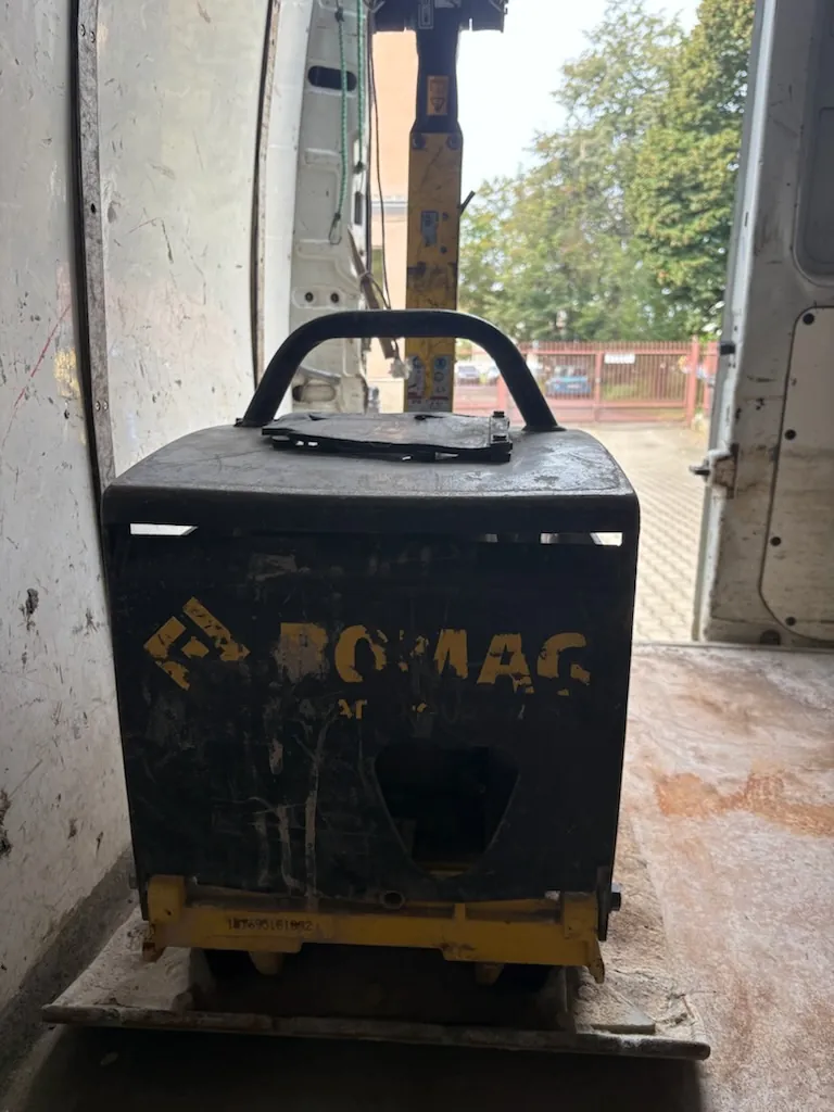 Czarna, metalowa zagęszczarka do gruntu z żółtym napisem 'BOMAG' na przednim panelu. Urządzenie posiada czarny uchwyt do przenoszenia na górze i żółtą podstawę. Maszyna znajduje się wewnątrz białego pojazdu dostawczego, w tle widoczny jest fragment żółtego dźwigu oraz zielone drzewa za ogrodzeniem.