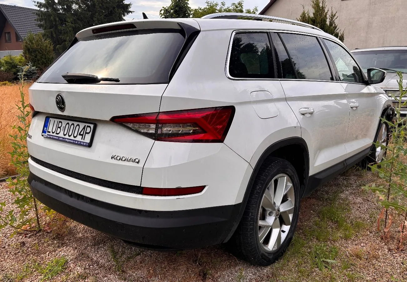 Biały samochód typu SUV marki Volkswagen zaparkowany na trawie. Widoczny jest tył i prawy bok pojazdu. Samochód posiada tablicę rejestracyjną o numerze LU 004P.