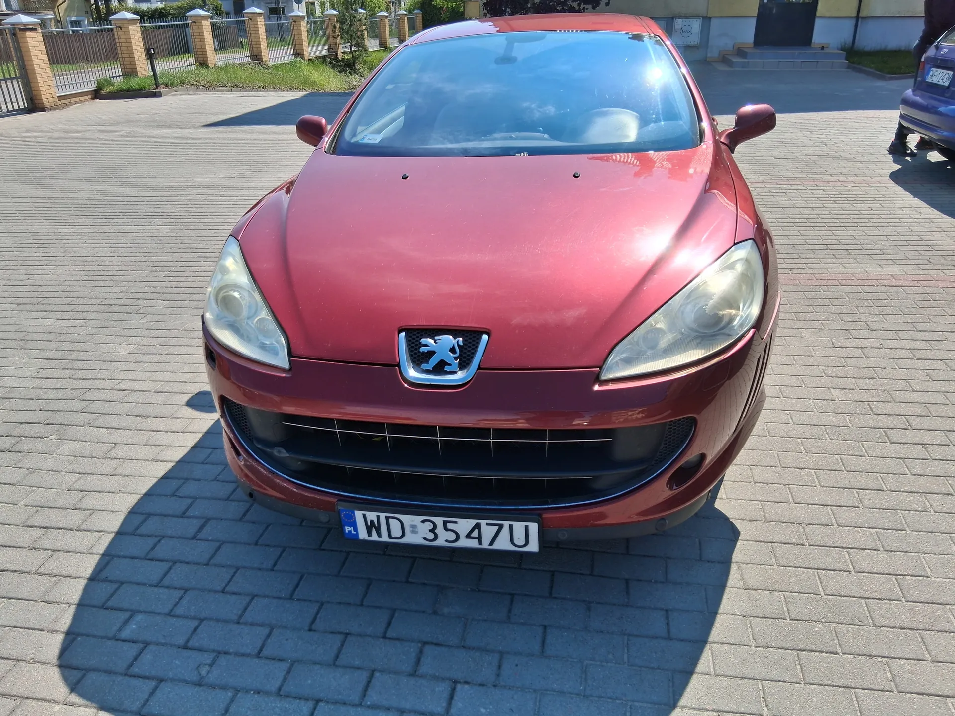 Czerwony samochód marki Peugeot z przodu, z widocznym logo lwa na masce. Tablica rejestracyjna z napisem WD 3547U oraz oznaczeniem PL po lewej stronie. Samochód stoi na brukowanej powierzchni, na tle ogrodzenia z ceglanymi filarami, zielonej trawy i częściowo widocznego innego pojazdu po prawej stronie. W tle budynki i drzewa.