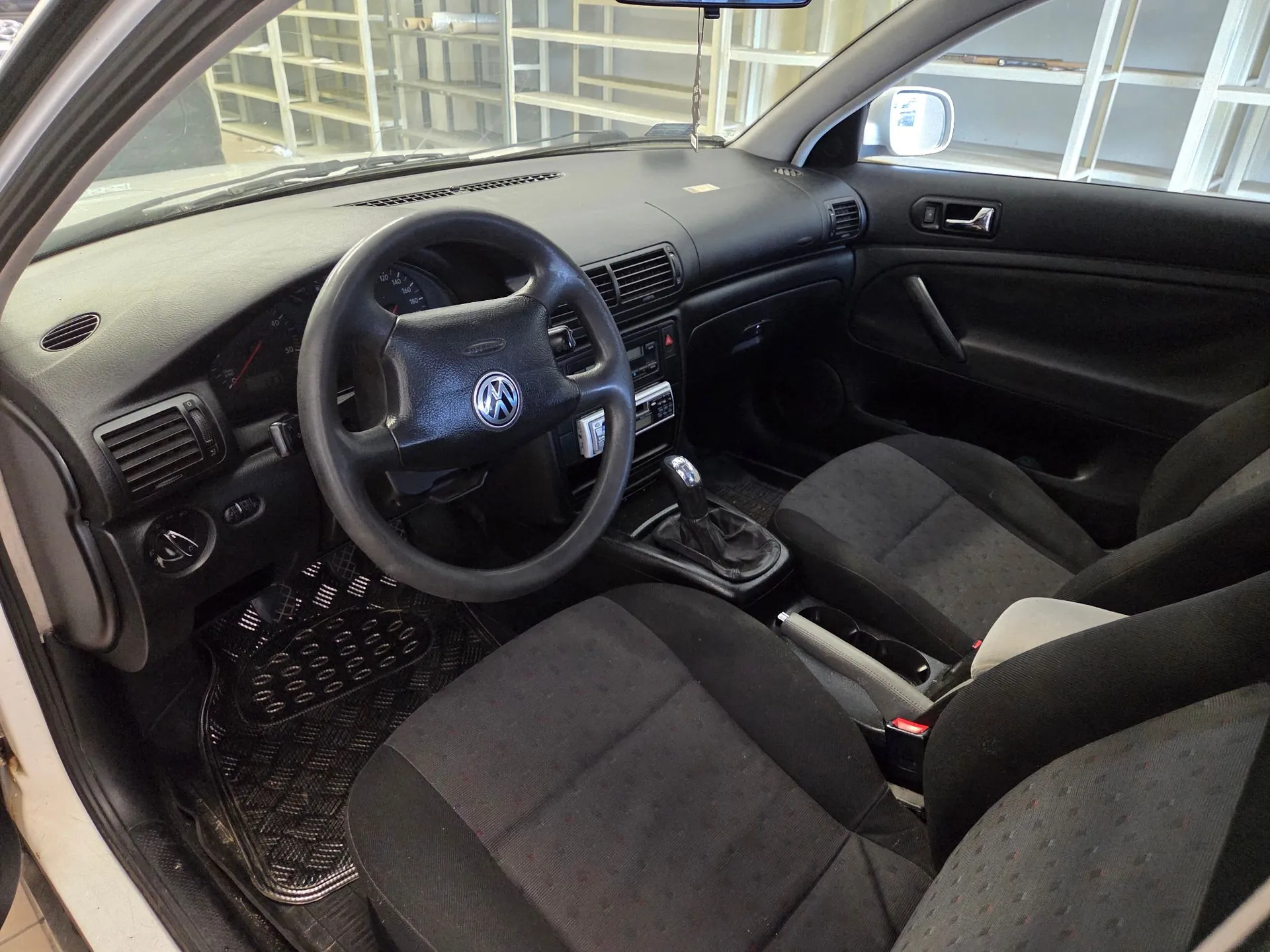 Wnętrze samochodu marki Volkswagen, widok z perspektywy pasażera na stronę kierowcy. Widoczna jest czarna deska rozdzielcza, czarna kierownica z logo VW w centrum, czarne materiałowe fotele oraz gumowe dywaniki z wypustkami. Samochód posiada manualną skrzynię biegów.