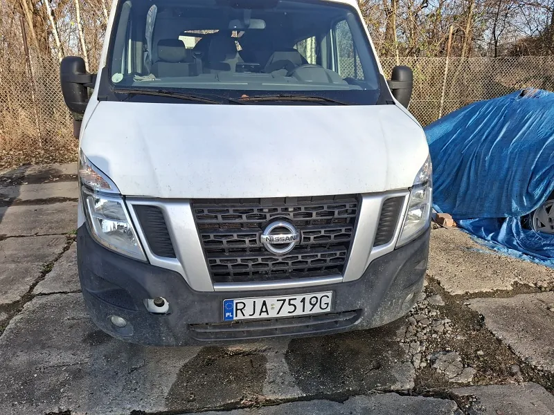 Biały samochód dostawczy marki Nissan z przodu, z widoczną tablicą rejestracyjną PL RJA 7519G. Samochód stoi na pękniętym betonie, w tle siatka ogrodzeniowa i suche drzewa. Po prawej stronie obrazu znajduje się przedmiot przykryty niebieską folią. Przednia część pojazdu jest lekko zabrudzona, a logo Nissan widoczne na atrapie chłodnicy.