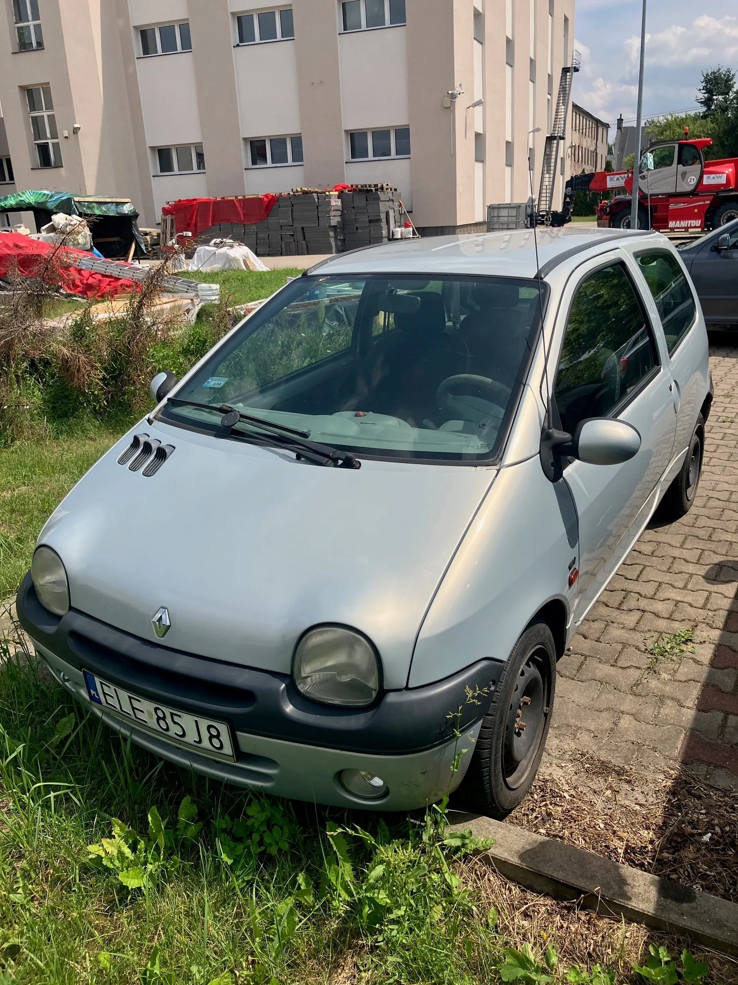 Srebrny samochód marki Renault Twingo zaparkowany na krawężniku obok trawnika. W tle widoczny jest jasny budynek mieszkalny z oknami oraz czerwony wózek widłowy marki Manitou. Samochód ma tablicę rejestracyjną o treści ELF 85J8.
