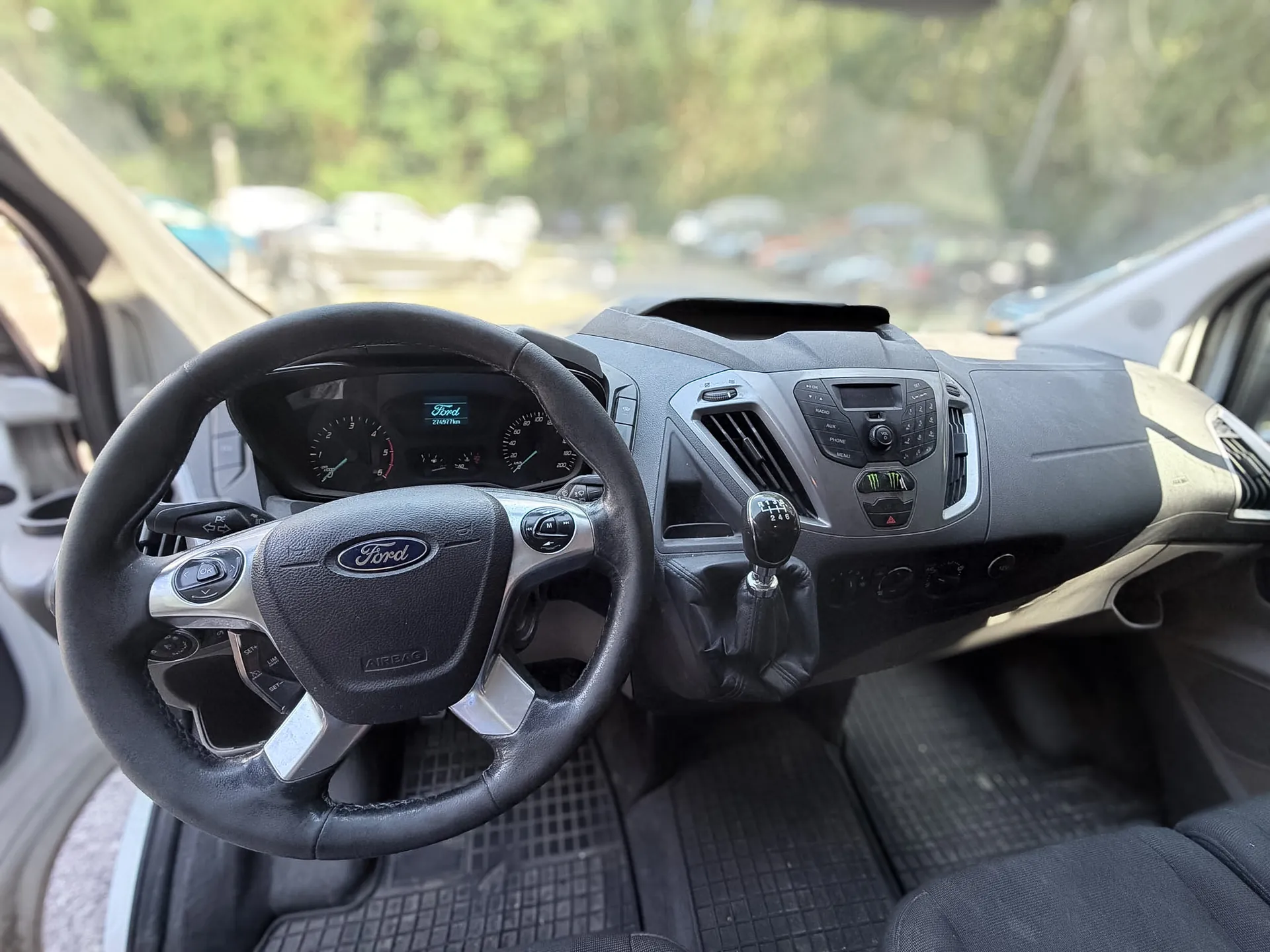 Wnętrze samochodu marki Ford widziane z perspektywy kierowcy. Widoczna jest czarna, skórzana kierownica z logo Ford na środku, deska rozdzielcza z zegarami, konsola centralna z radiem i przyciskami oraz dźwignia zmiany biegów. W tle, za przednią szybą, widać rozmyty parking z samochodami i zieleń drzew.