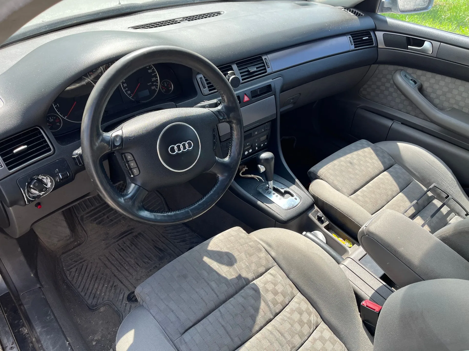 Wnętrze samochodu marki Audi widziane z perspektywy pasażera. Widoczna jest czarna kierownica z logo Audi, deska rozdzielcza z zegarami, konsola środkowa z dźwignią zmiany biegów oraz dwa przednie fotele obite szarym materiałem w delikatną kratkę.