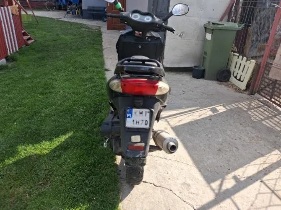 Z tyłu widoczny czarny skuter z tablicą rejestracyjną KM1 1H70 (z niebieskim pasem z napisem PL po lewej stronie). Skuter stoi na betonowej powierzchni, po lewej stronie trawa, po prawej zielony pojemnik na śmieci, biały płotek i metalowa brama. W tle budynek z białą ścianą oraz rower i niebieski pojemnik. Skuter posiada czerwone światło tylnie, wydech i czarny kufer na bagaż.