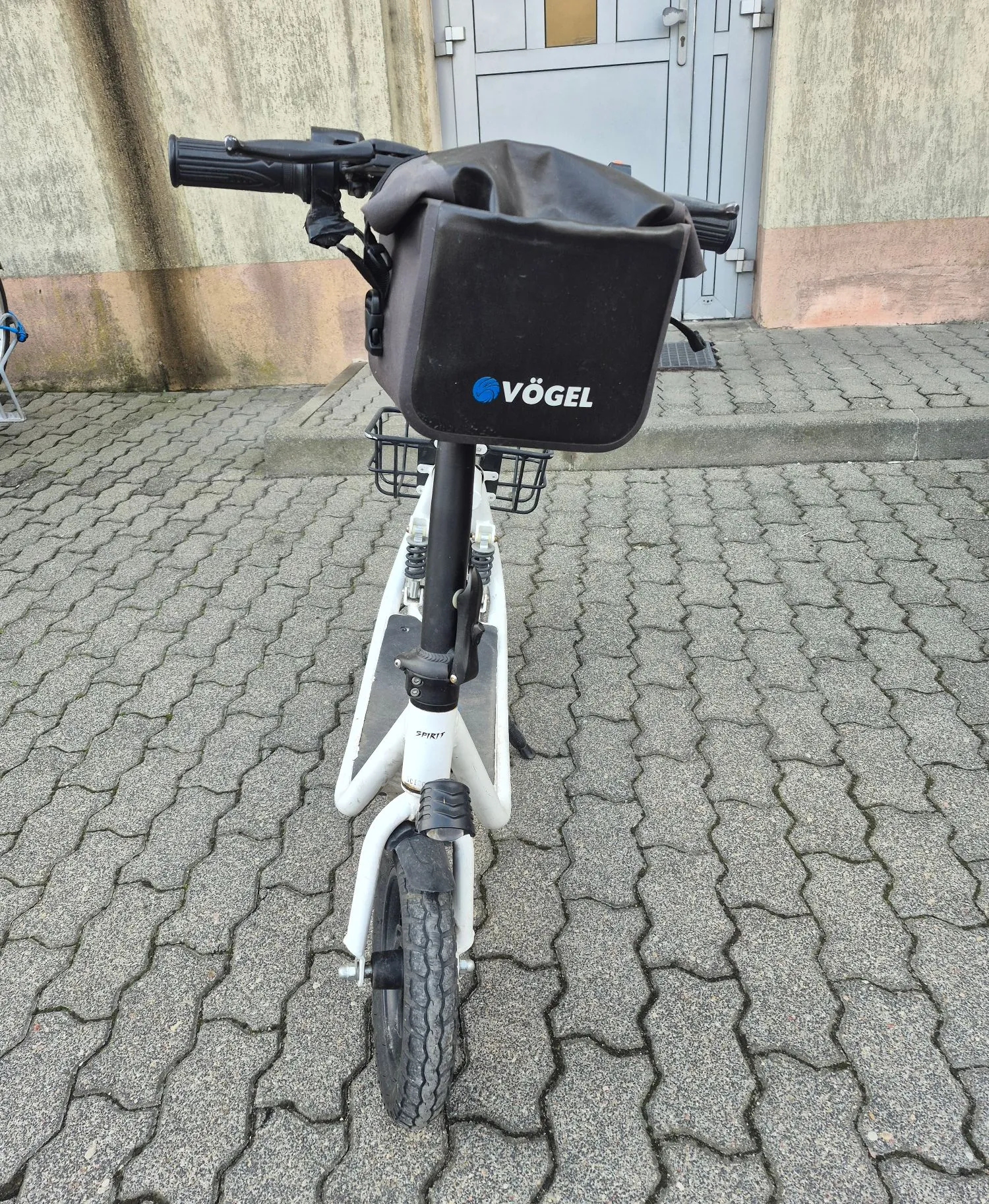 Biała hulajnoga elektryczna z czarną torbą zamontowaną na kierownicy. Na torbie widnieje niebiesko-biały napis 'VÖGEL'. Hulajnoga stoi na szarym, kostkowym chodniku przed jasną ścianą budynku z szarymi drzwiami.