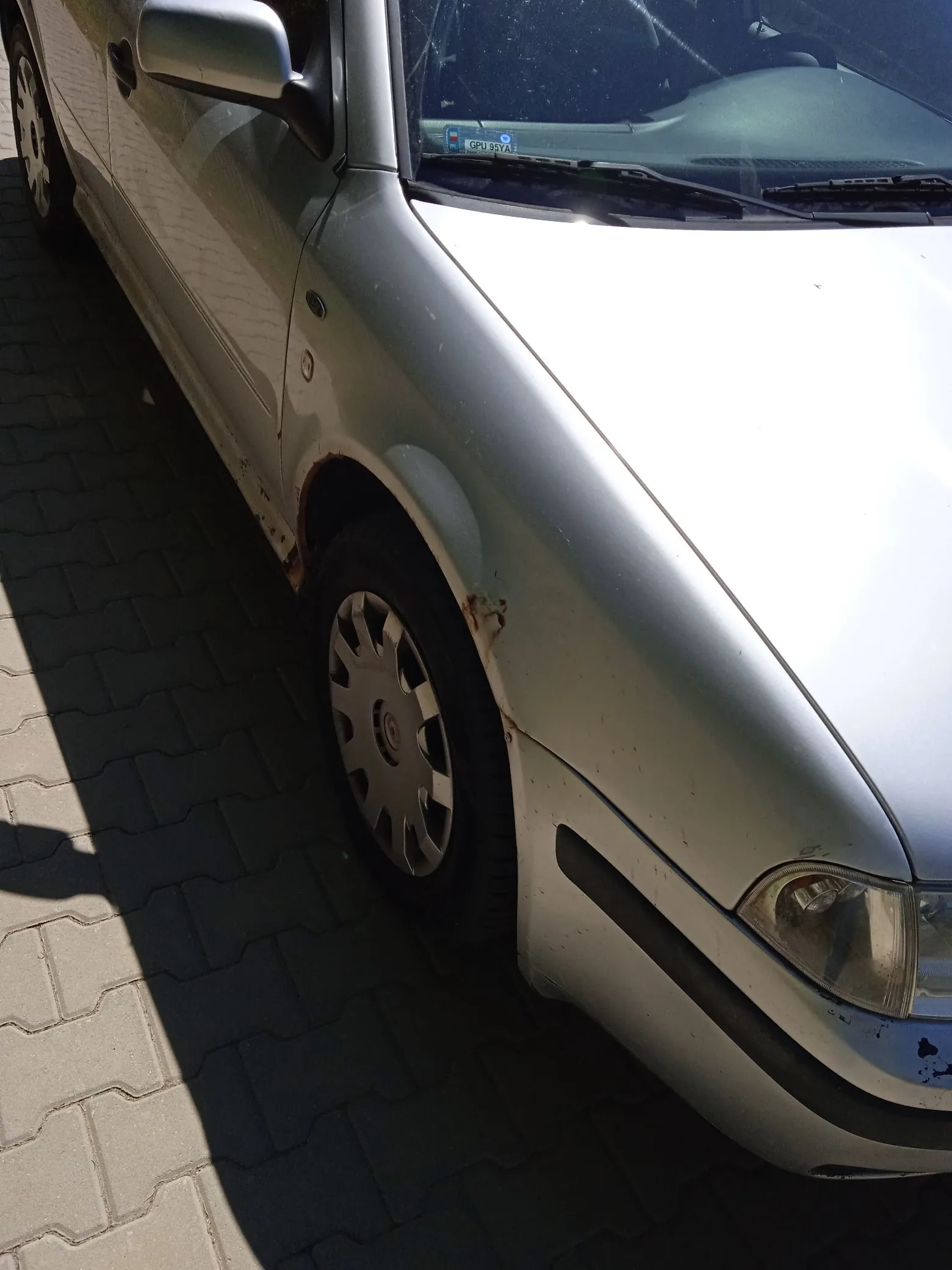 Zbliżenie na przedni prawy błotnik i koło srebrnego samochodu osobowego. Na nadkolu widoczne są wyraźne ślady korozji i rdzy. Samochód zaparkowany jest na kostce brukowej w pełnym słońcu, co tworzy ostry cień po lewej stronie obrazu.