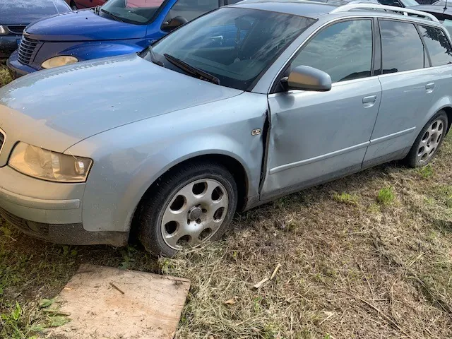 Srebrny samochód typu kombi zaparkowany na trawiastym terenie. Pojazd posiada widoczne uszkodzenia karoserii w postaci wgniecenia na przednich drzwiach kierowcy oraz na błotniku nad przednim kołem. W tle widoczny jest fragment niebieskiego samochodu.
