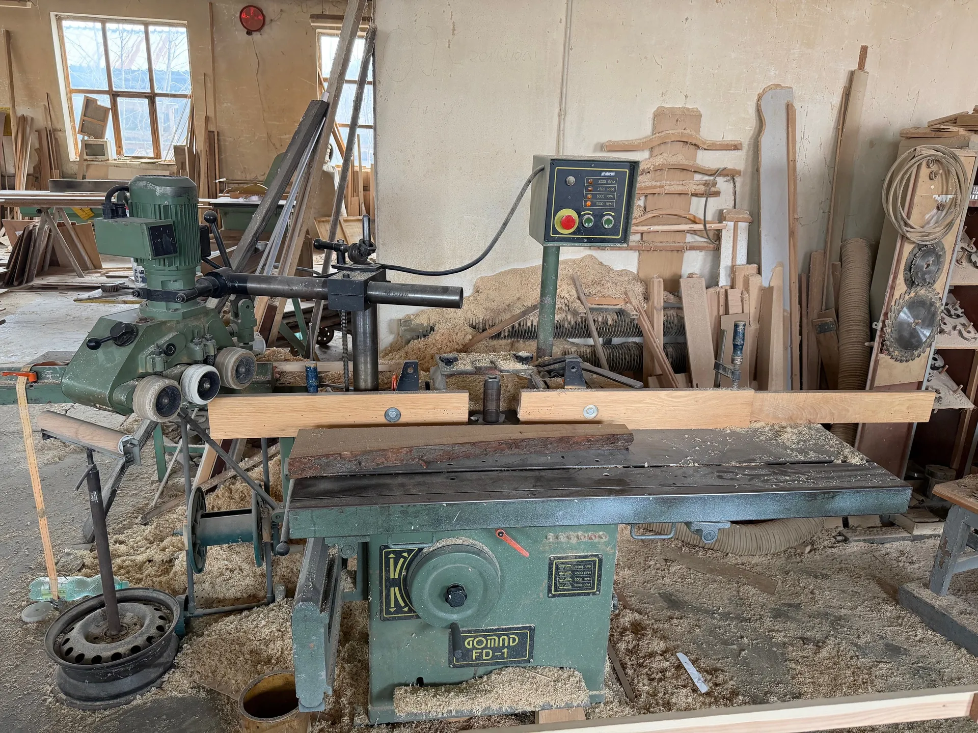 Wnętrze warsztatu stolarskiego z zielonym maszyną do obróbki drewna oznaczoną napisem GOMAD FD-1. Na maszynie widoczny panel sterowania z czerwonym przyciskiem i wyświetlaczem. Podłoga pokryta jest wiórami, a wokół znajdują się kawałki drewna, deski i narzędzia. W tle okna z widokiem na zewnątrz, czerwony alarm pożarowy na ścianie oraz różne materiały i urządzenia warsztatowe.