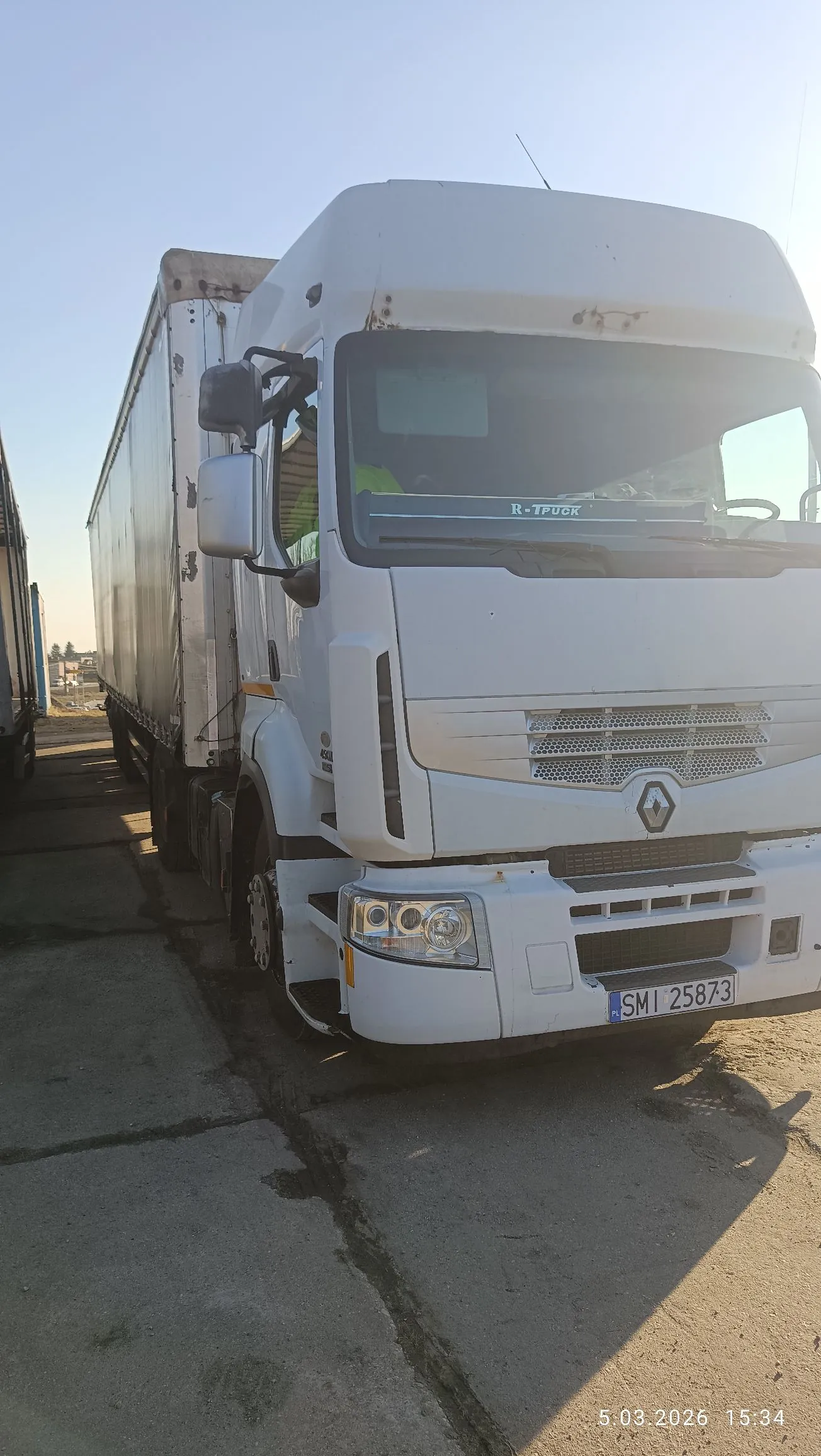 Biała ciężarówka marki Renault z naczepą, zaparkowana na betonowym placu. Na przedniej szybie widnieje napis 'R-TRUCK'. Pojazd posiada polską tablicę rejestracyjną o numerze SMI 25873. W prawym dolnym rogu zdjęcia znajduje się data i godzina: 5.03.2026 15:34.