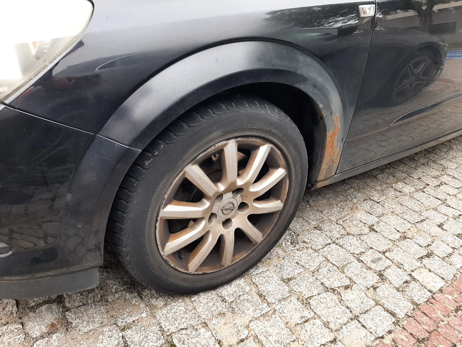 Zbliżenie na przednie lewe koło czarnego samochodu zaparkowanego na kostce brukowej. Felga aluminiowa jest zabrudzona pyłem z klocków hamulcowych i posiada logo Nissana w centrum. Na nadkolu, tuż za kołem, widoczne jest wyraźne ognisko korozji z odpryskiem lakieru.