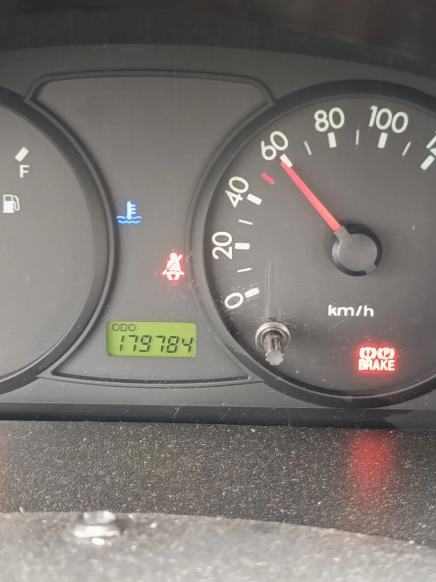Zbliżenie na deskę rozdzielczą samochodu. Widoczny jest prędkościomierz z zaznaczoną prędkością około 65 km/h, wskaźnik temperatury cieczy chłodzącej (niebieska ikona), kontrolka niezapiętych pasów bezpieczeństwa (czerwona ikona) oraz kontrolka hamulca ręcznego z napisem 'BRAKE'. Cyfrowy licznik przebiegu (ODO) wskazuje wartość 179784.