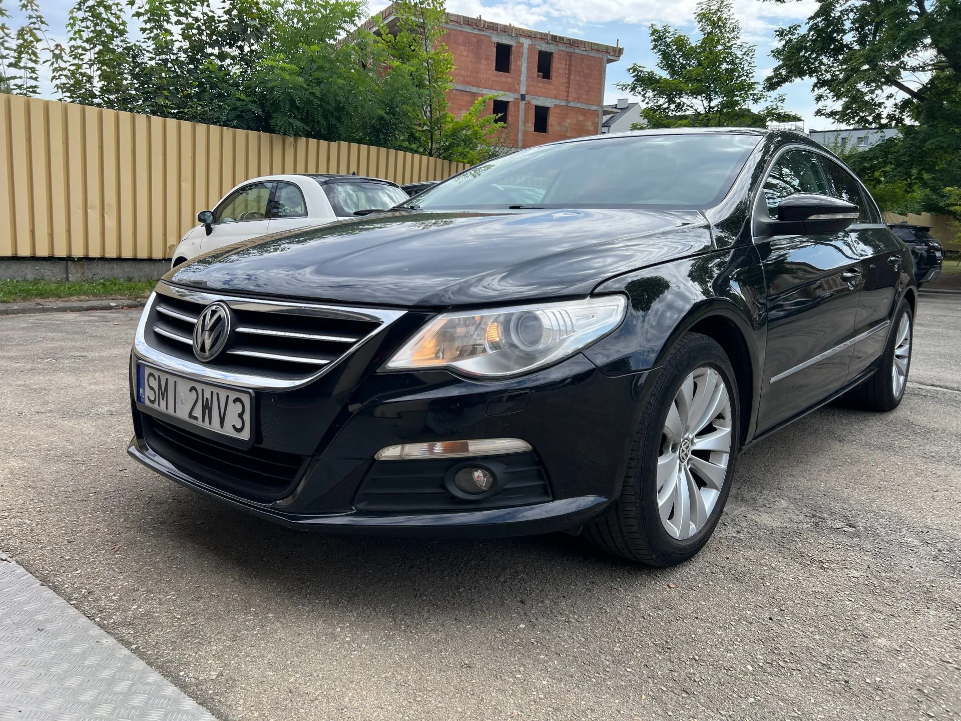Czarny samochód osobowy marki Volkswagen zaparkowany na asfaltowym podjeździe. Widoczna jest przednia część pojazdu z rejestracją SMI 2WV3. W tle znajduje się biały samochód, żółty płot oraz budynek w stanie surowym z czerwonej cegły.