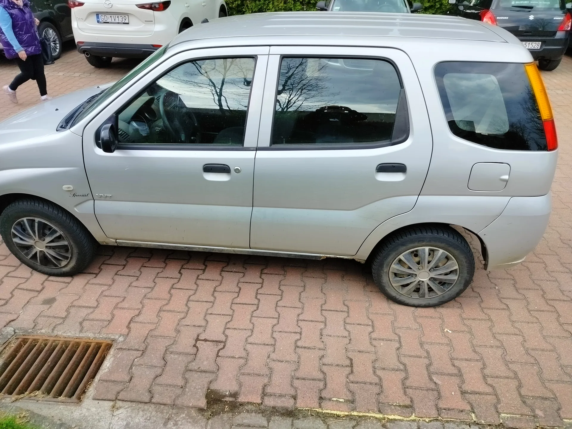 Zdjęcie przedstawia srebrny samochód osobowy typu hatchback zaparkowany na brukowanej nawierzchni. W tle widoczne są inne pojazdy: biały Mazda z tablicą rejestracyjną GD 1V139 oraz czarny Suzuki z tablicą 51528. Po lewej stronie obrazu przechodzi osoba w fioletowej kamizelce i czarnych spodniach. Na pierwszym planie znajduje się kanał odpływowy. Odbicia drzew widoczne są w szybach samochodu. Niebo jest zachmurzone.
