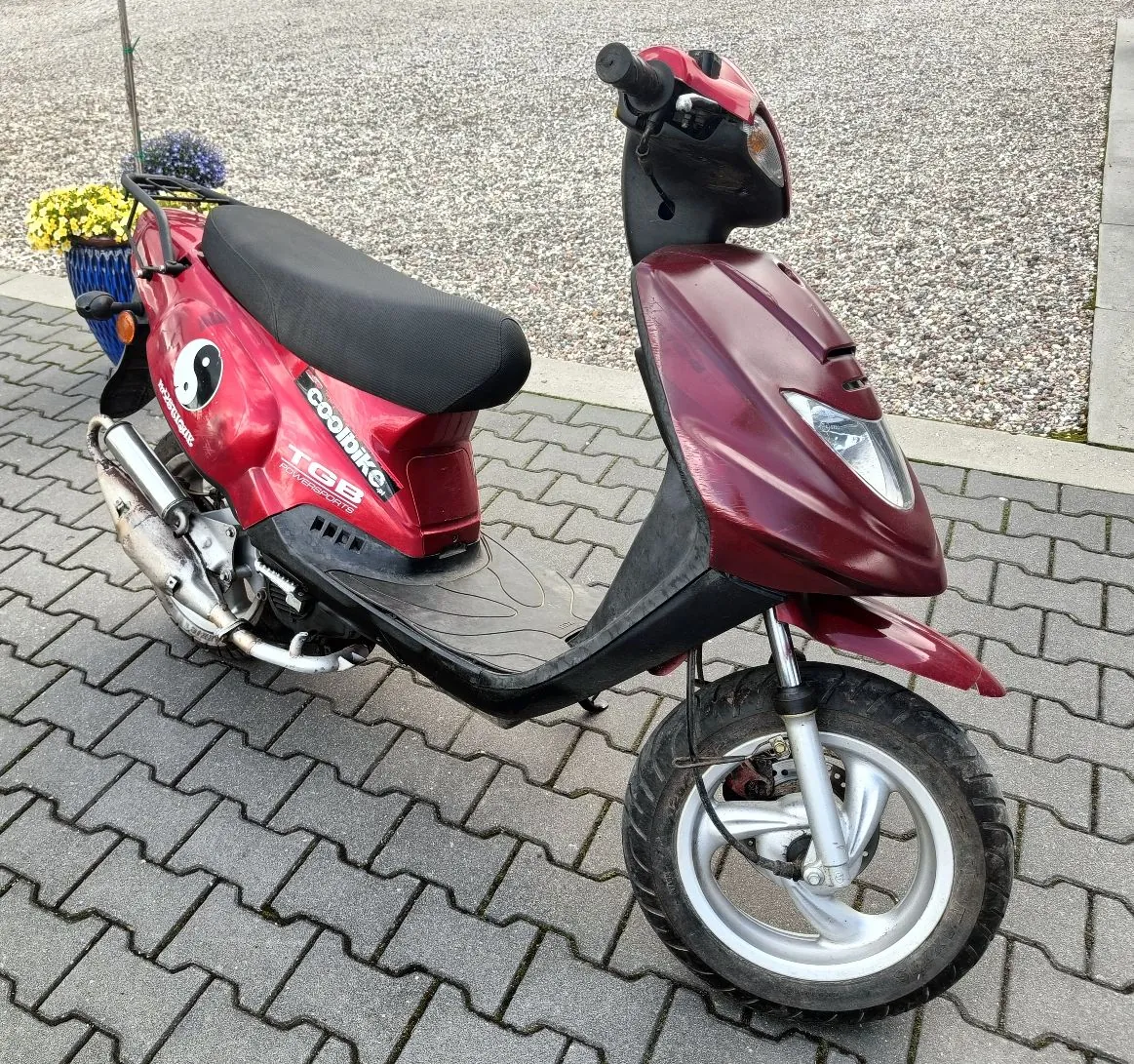 Czerwony skuter marki TGB Coolbike zaparkowany na szarym chodniku z kostki brukowej. Na boku skutera widnieje biały symbol yin-yang oraz napisy 'coolbike' i 'TGB'. W tle widać żwirową nawierzchnię oraz niebieską donicę z żółtymi kwiatami.