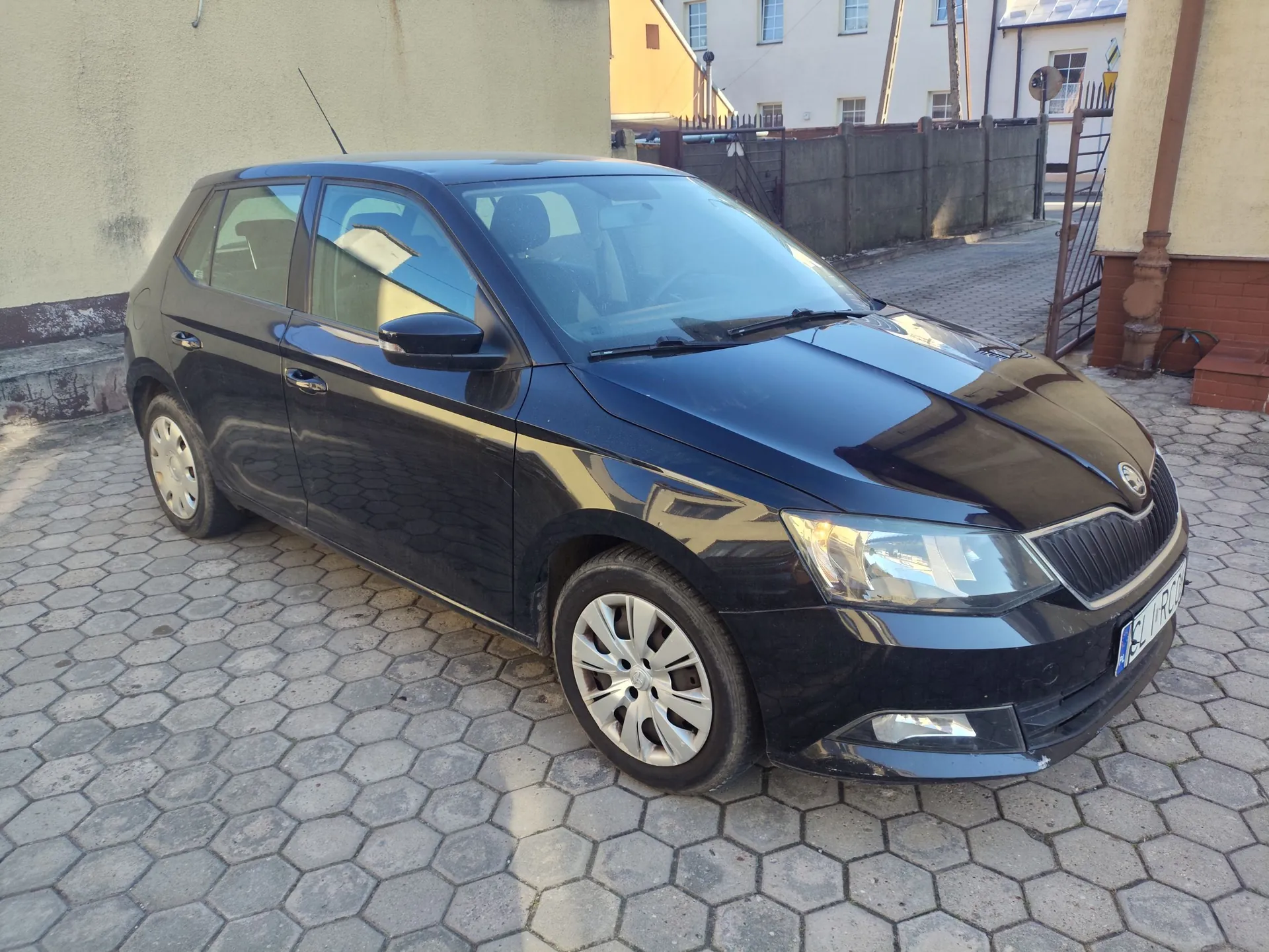 Czarny samochód osobowy marki Skoda zaparkowany na podjeździe z szarej kostki brukowej o kształcie sześcioboków. Auto jest ustawione pod kątem, widoczny jest przód i lewy bok pojazdu. W tle znajduje się jasna ściana budynku oraz ogrodzenie z ciemnego betonu.