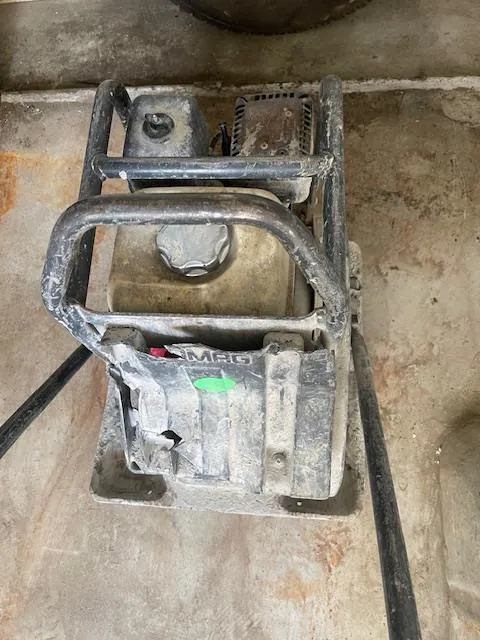 Zdjęcie przedstawia brudny, metalowy sprzęt przemysłowy, prawdopodobnie wibracyjny zagęszczacz betonu. Urządzenie posiada metalowe uchwyty, zbiornik paliwa z pokrętłem, zielony naklejkę oraz widoczny napis 'MPC' na przedniej części. Tło stanowi betonowa podłoga z plamami i śladami zużycia. Całość pokryta jest warstwą kurzu i brudu.