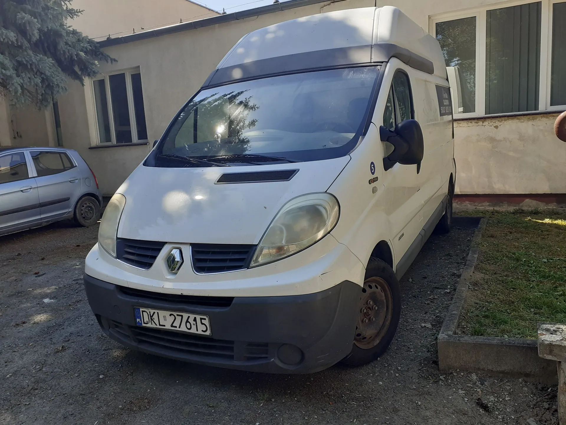 Biały samochód dostawczy marki Renault zaparkowany na podjeździe przed jasnym budynkiem. Pojazd ma wysoką nadbudowę dachu i polską tablicę rejestracyjną o numerze DKL 27615. W tle po lewej stronie widoczny jest fragment srebrnego samochodu osobowego oraz zielone drzewo.