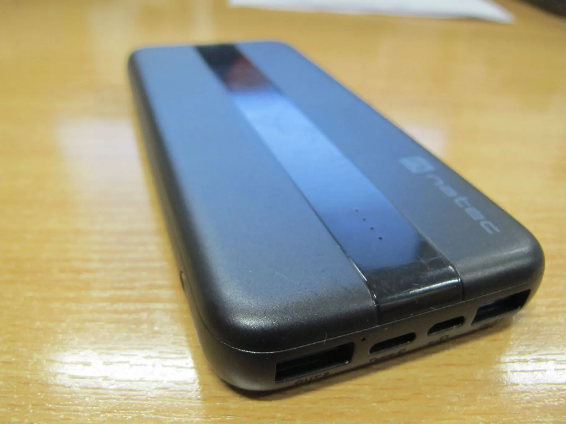 Czarny powerbank marki natec leżący na drewnianej powierzchni. Urządzenie ma podłużny kształt z zaokrąglonymi krawędziami, błyszczący pas na górnej obudowie oraz widoczne porty wejścia/wyjścia na jednym z krótszych boków.