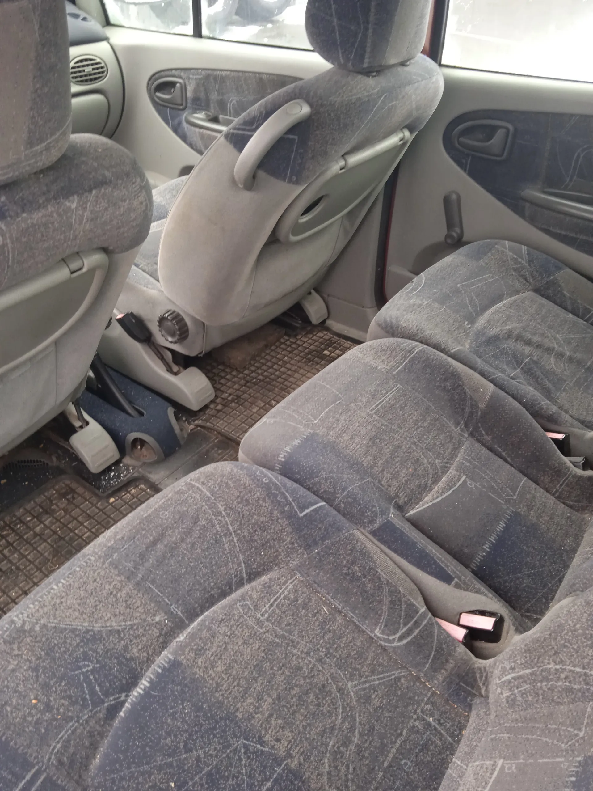 Wnętrze samochodu z widokiem na przód. Widoczne są zanieczyszczone siedzenia pasażerskie i kierowcy w szaro-niebieskim wzorze, podłoga z matami, pedały, dźwignia zmiany biegów oraz fragmenty drzwi z uchwytami i oknami. W tle przez przednią szybę widać zewnętrzne otoczenie.