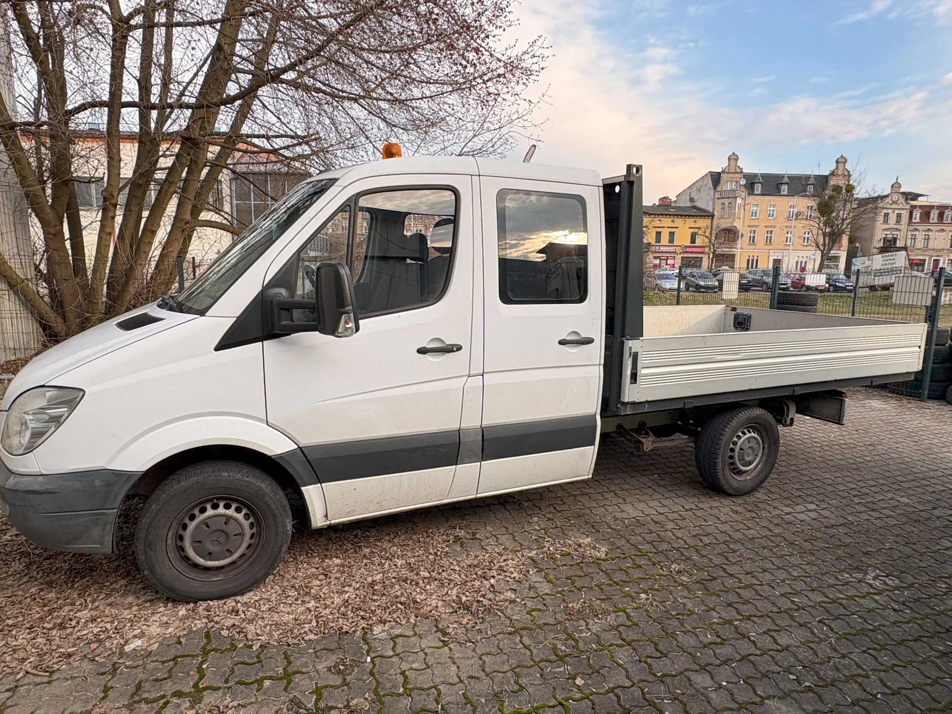 Biały samochód dostawczy z platformą załadowczą zaparkowany na brukowanej powierzchni. Pojazd posiada czarną listwę boczną, pomarańczowe światło na dachu i czarne lusterko boczne. W tle widoczne są drzewa bez liści, budynki mieszkalne z klasyczną architekturą, parkujące samochody oraz niebo z rozproszonymi chmurami. Platforma załadowcza jest pusta, a na ziemi leżą suche liście.