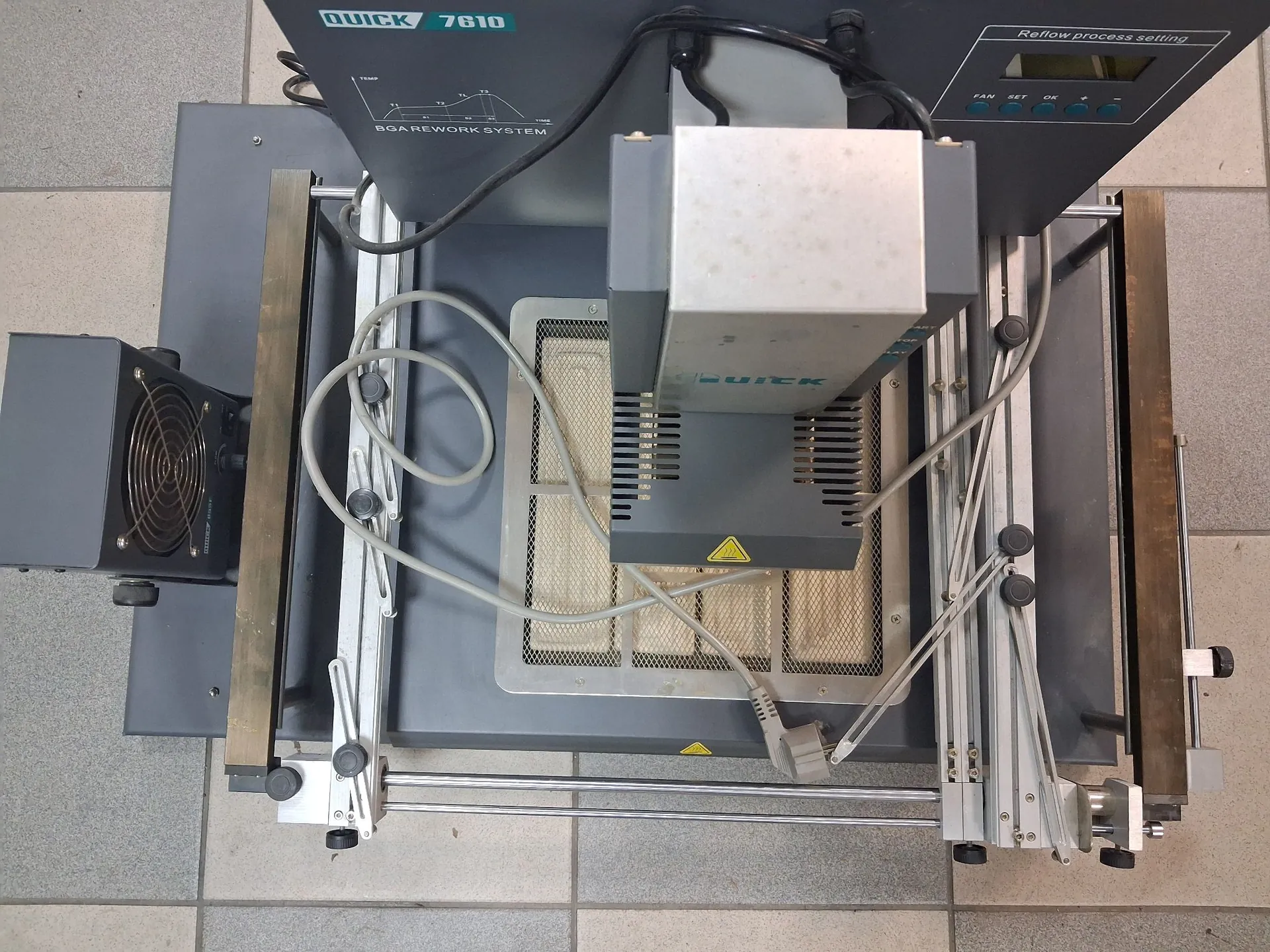 Widok z góry na stację do reballingu BGA marki QUICK, model 7610. Urządzenie składa się z szarej podstawy, na której zamontowany jest system prowadnic, grzałka dolna w formie perforowanej płyty oraz ruchoma głowica grzewcza z logo QUICK. W górnej części znajduje się panel sterowania z napisem 'Reflow process setting' oraz wyświetlaczem. Po lewej stronie widoczny jest zewnętrzny moduł chłodzący z wentylatorem. Całość stoi na jasnej, kafelkowej podłodze.