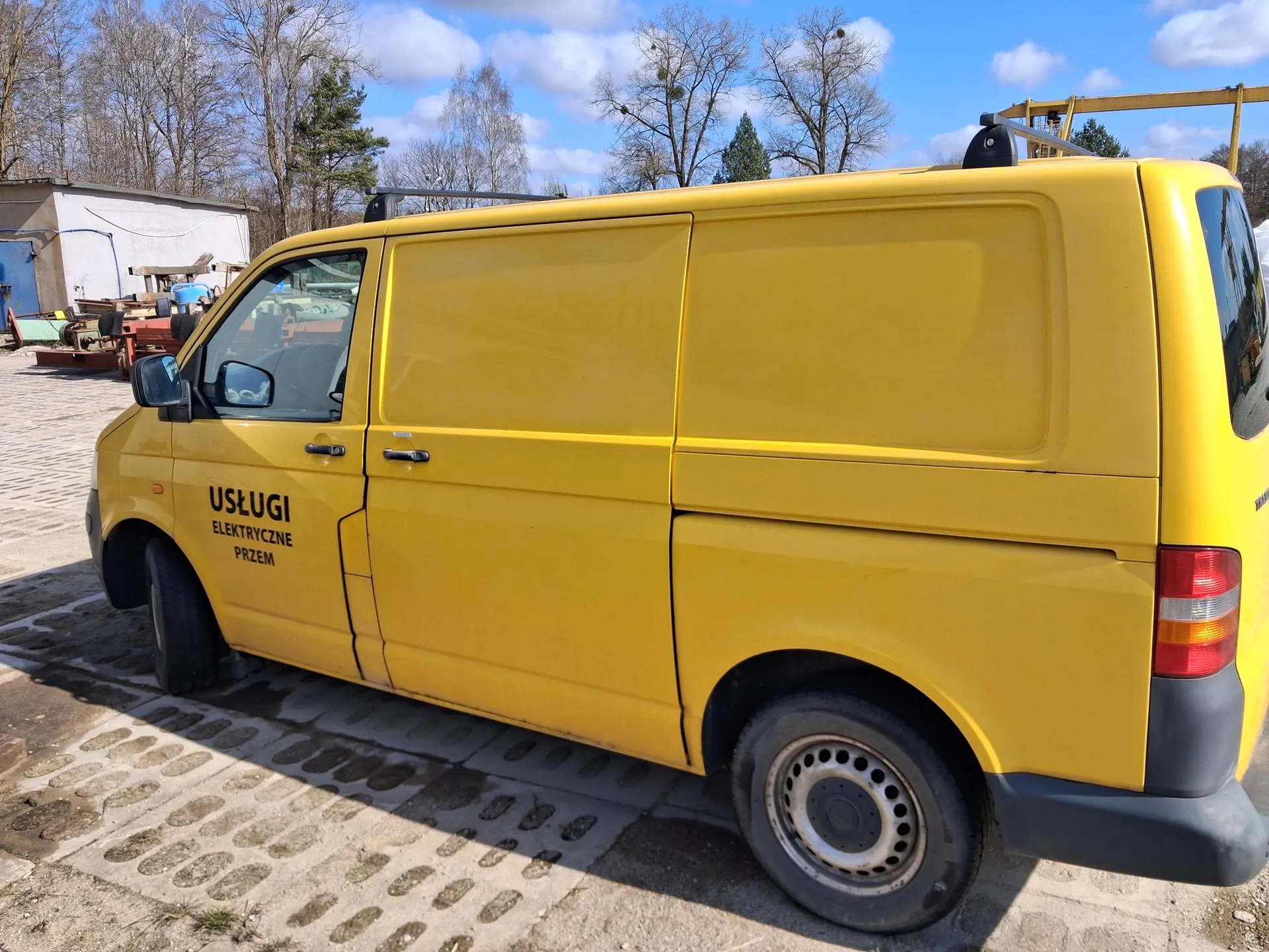 Żółty samochód dostawczy zaparkowany na brukowanej powierzchni. Na boku pojazdu widoczny czarny napis: USŁUGI ELEKTRYCZNE PRZEM. W tle drzewa bez liści, biały budynek, maszyny i niebo z chmurami. Samochód ma czarne felgi i czerwone światła tylna.