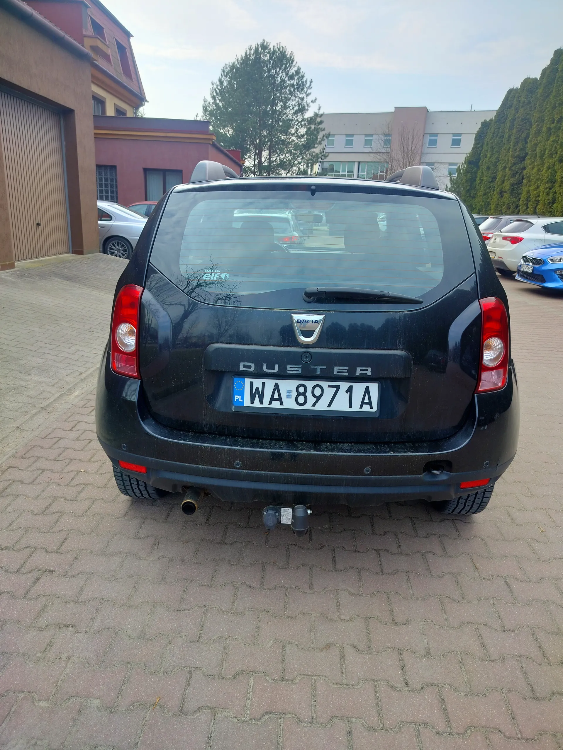 Tył czarnego samochodu Dacia Duster z tablicą rejestracyjną WA 8971A (PL) zaparkowanego na brukowanej powierzchni. Widoczne są czerwone światła tylne, logo marki, napis DUSTER oraz naklejka na szybie. W tle budynki, drzewa i inne zaparkowane pojazdy.