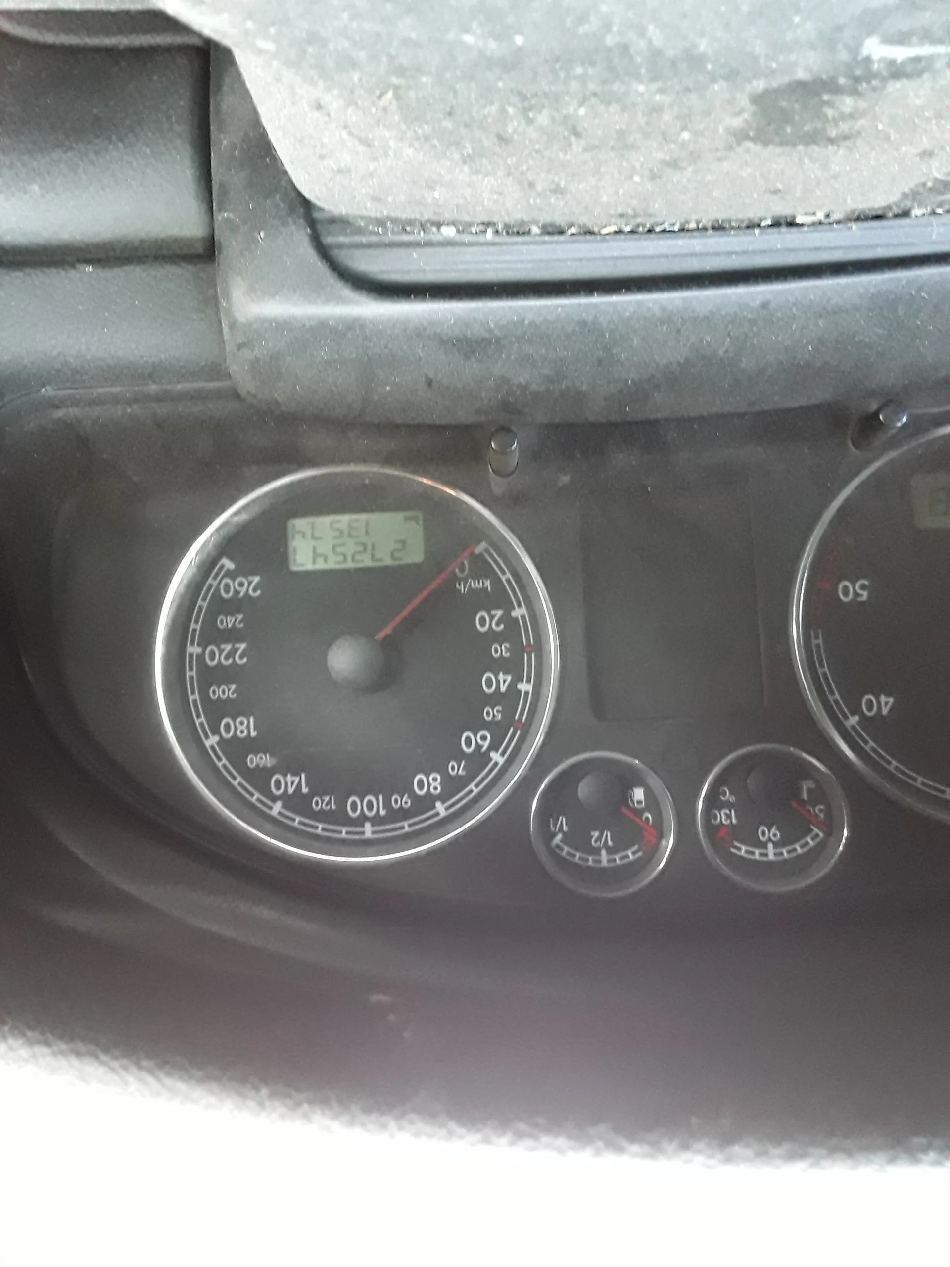 Zbliżenie na deski rozdzielcze samochodu. Wielki wskaźnik prędkości z zakresem 0-260 km/h, igła wskazuje 0 km/h. Cyfrowy wyświetlacz pokazuje wartości '27254' i '135.7'. Poniżej znajdują się dwa mniejsze wskaźniki: paliwa (z podziałką 1/2) i temperatury chłodniczki (do 130°C). Widoczny fragment tachometru z zakresem do 50.