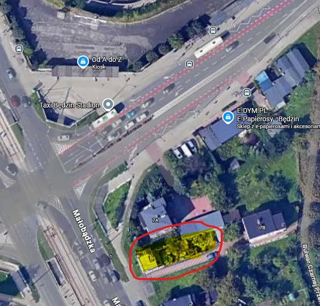 Zrzut ekranu z mapy satelitarnej przedstawiający skrzyżowanie ulic, w tym ulicę Małobadzką. Na obrazie widoczne są budynki, drogi z pasami ruchu oraz zieleń miejska. Na mapie znajdują się znaczniki punktów usługowych z napisami: 'Od A do Z Kiosk', 'Taxi Będzin Stadion' oraz 'E-DYM.PL - E-Papierosy - Będzin'. Jeden z budynków w dolnej części obrazu jest zaznaczony grubą czerwoną linią, a jego dach wypełniony jest jaskrawym żółtym kolorem.