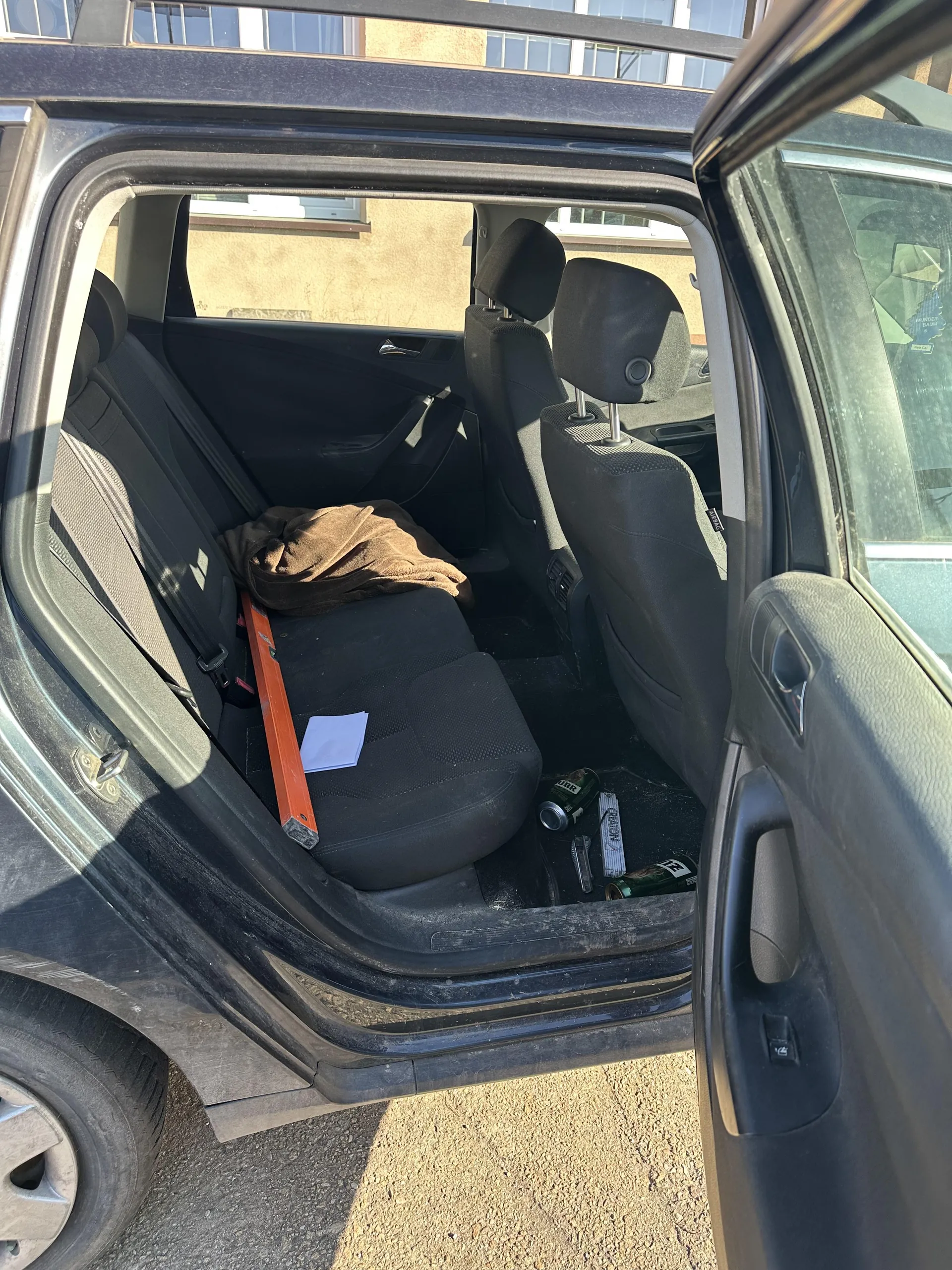 Wnętrze czarnego samochodu z otwartymi tylnymi drzwiami. Widoczne czarne siedzenia, na tylnym siedzeniu brązowy ręcznik lub koc, pomarańczowy poziomnik i biały arkusz papieru. Na podłodze znajdują się puste butelki, puszka oraz paczka papierosów z częściowo czytelnym napisem. W tle budynki z oknami, a pod samochodem asfalt. Drzwi samochodu są otwarte, a wnętrze wydaje się nieporządne.