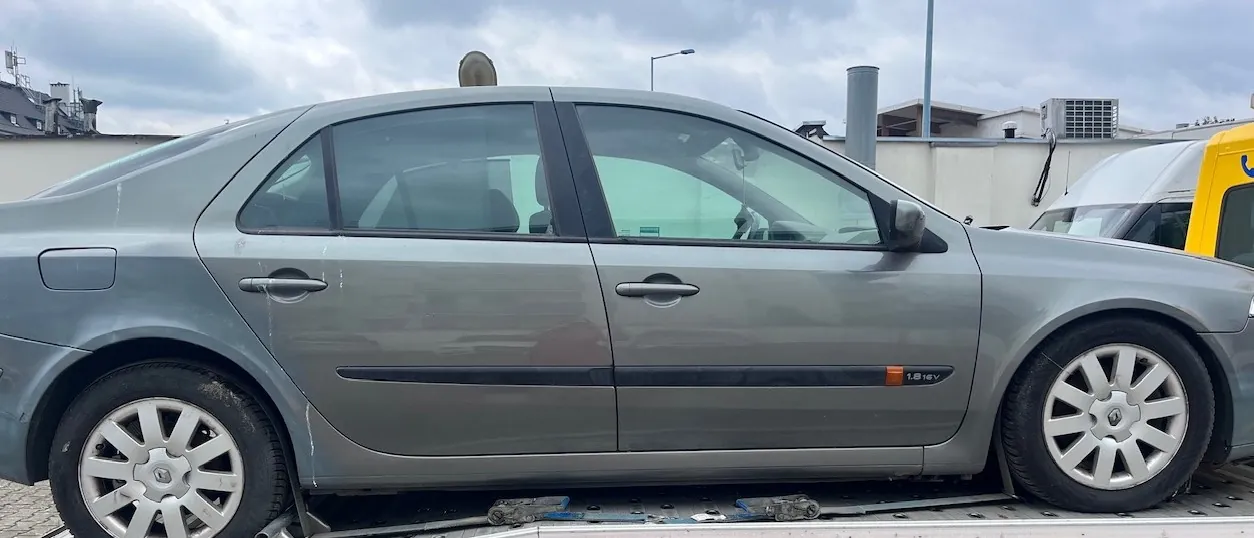 Widok z boku szarego samochodu osobowego typu sedan, zaparkowanego na platformie lawety. Pojazd ma pięcioramienne felgi aluminiowe i czarną listwę ochronną na drzwiach z napisem '1.8 16V'. W tle widoczne jest zachmurzone niebo oraz fragmenty budynków przemysłowych.