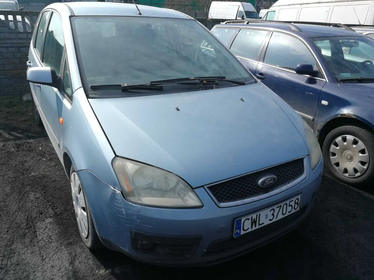 Przedni widok jasnoniebieskiego samochodu marki Ford z tablicą rejestracyjną PL CWL 37058. Po prawej stronie widoczny jest ciemnoniebieski samochód. W tle znajdują się białe furgony, kamienna ściana oraz zielone elementy otoczenia. Samochody zaparkowane są na asfaltowej powierzchni.