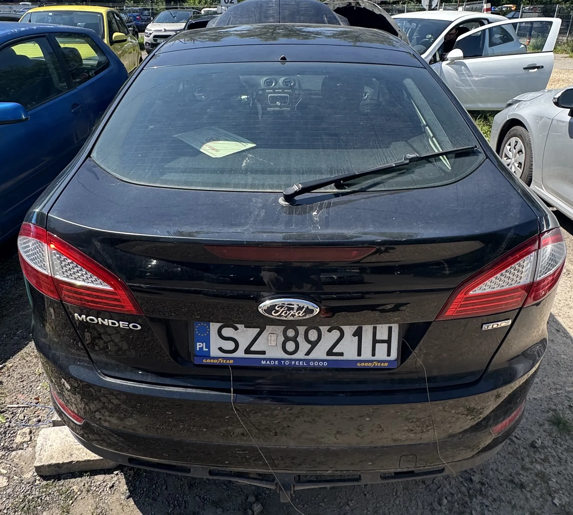 Tylny widok czarnego samochodu marki Ford Mondeo z polską tablicą rejestracyjną o numerze SZ 8921H. Samochód znajduje się na parkingu obok innych pojazdów, w tym niebieskiego i białego auta. Na tylnej szybie widoczny jest wycieraczka oraz kartka papieru. Na klapie bagażnika widnieją napisy 'Ford', 'MONDEO' oraz 'TDC'.