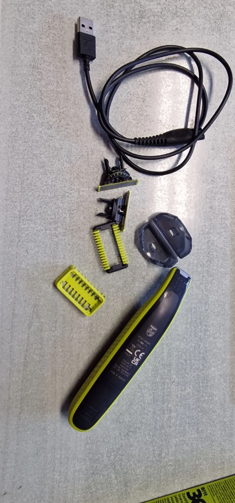 Zestaw do pielęgnacji zarostu ułożony na szarym, teksturowanym tle. W skład zestawu wchodzi: czarny kabel USB, trzy wymienne nasadki do golarki (jedna z metalowym ostrzem, jedna czarna plastikowa oraz dwie jasnozielone grzebienie o różnej długości), przezroczysta plastikowa osłona oraz główna jednostka trymera w kolorze czarnym z jasnozielonymi elementami. Na obudowie trymera widoczne są oznaczenia techniczne oraz logo marki.
