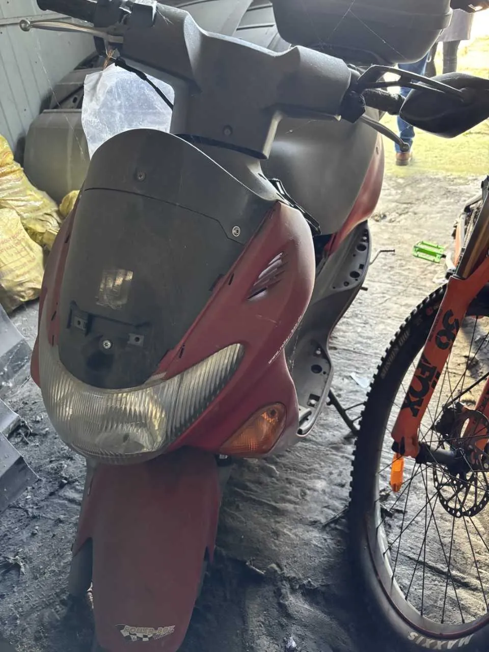 Czerwony skuter z czarnym wiatrochronem, reflektorem i kierunkowskazem. Po prawej stronie pomarańczowy rower z widocznymi napisami 'FOX' na ramie i 'MAXXIS' na oponie. W tle żółte worki, betonowa podłoga oraz osoba w niebieskich spodniach i brązowych butach. Na przednim błotniku skutera widoczny niewyraźny napis.