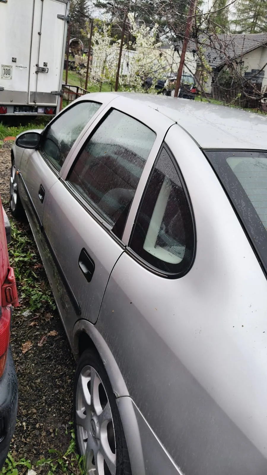 Srebrny samochód osobowy typu sedan zaparkowany na podwórku z drobnych kamieni i trawy. Na szybach widoczne krople wody. W tle biały przyczepa z napisem '100', drzewa z białymi kwiatami, dom z ciemnym dachem oraz fragment czerwonego samochodu po lewej stronie.