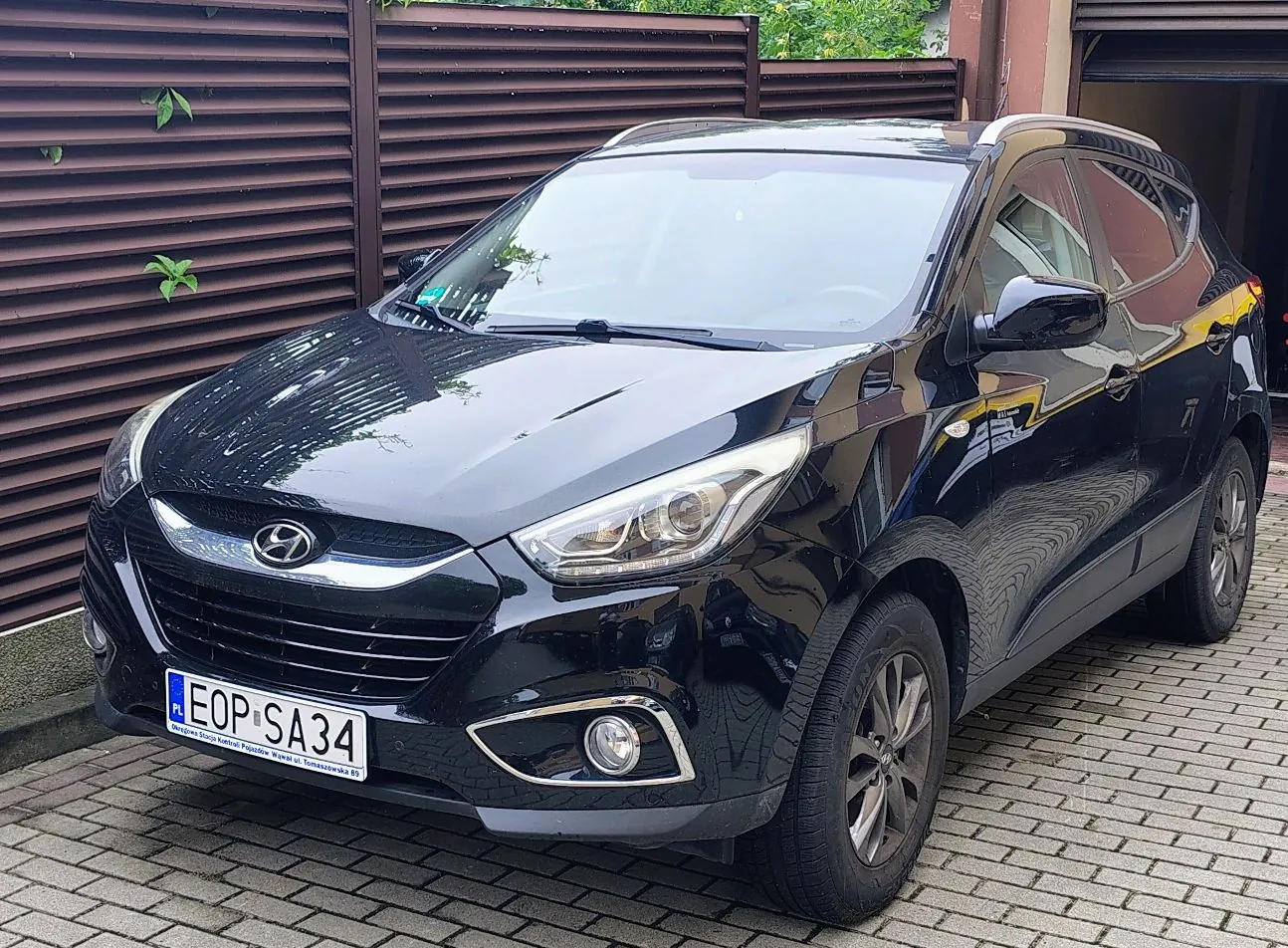 Czarny samochód marki Hyundai zaparkowany na kostce brukowej przed brązowym ogrodzeniem z poziomych listew. Pojazd posiada polską tablicę rejestracyjną o numerze EOP SA34.
