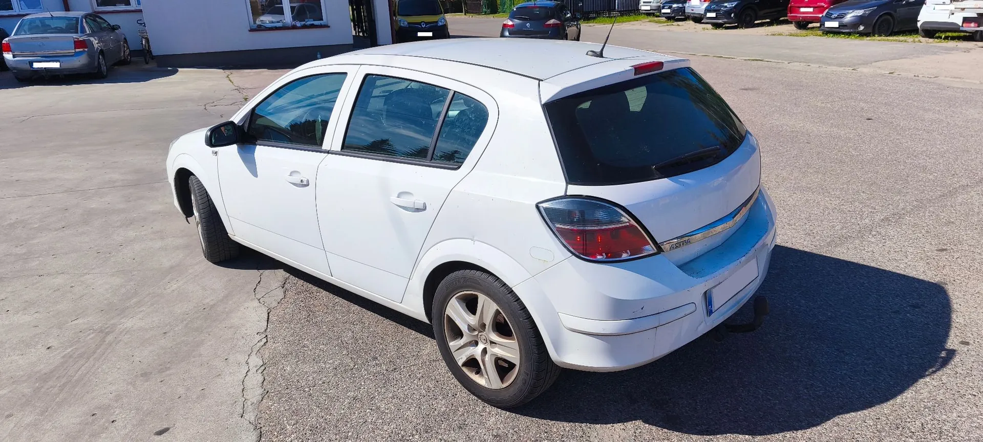 Biały samochód marki Opel Astra w nadwoziu hatchback, zaparkowany na asfaltowym placu. Pojazd jest pokazany z perspektywy tylno-bocznej od strony kierowcy. W tle widoczny jest fragment jasnego budynku oraz inne zaparkowane samochody.